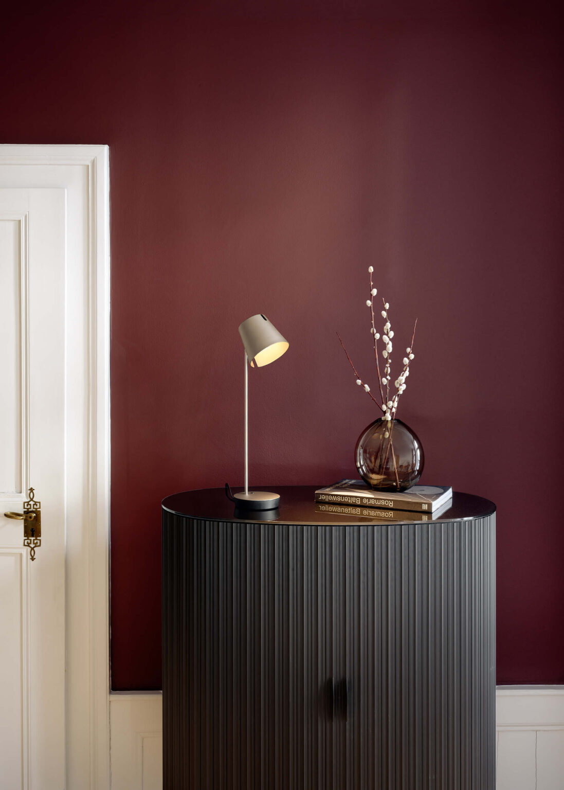 Baltensweiler Leuchte auf schwarzem Sideboard vor bordeauxfarbener Wand, mit Vase und Büchern bei teo jakob.