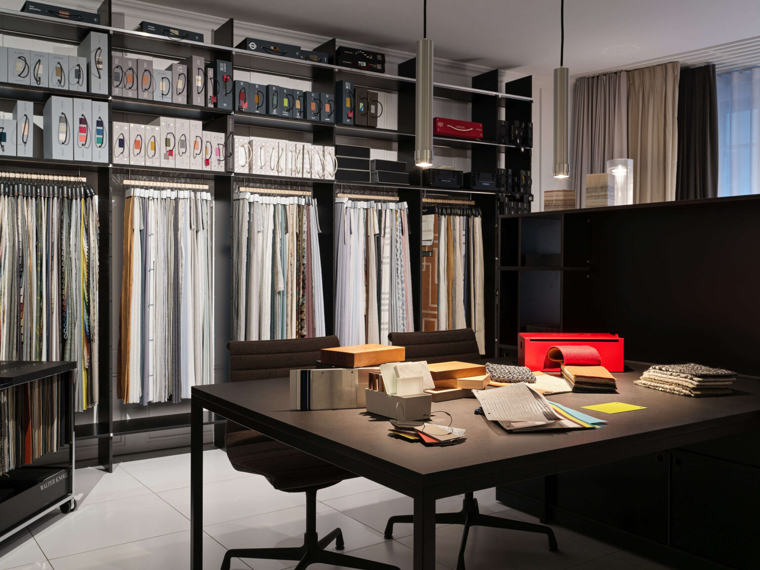 Showroom teo jakob Bern, Materialbibliothek mit hohen Regalen voller Stoffmuster und Boxen, davor ein grosser Tisch mit Materialproben und Leuchten über der Arbeitsfläche.
