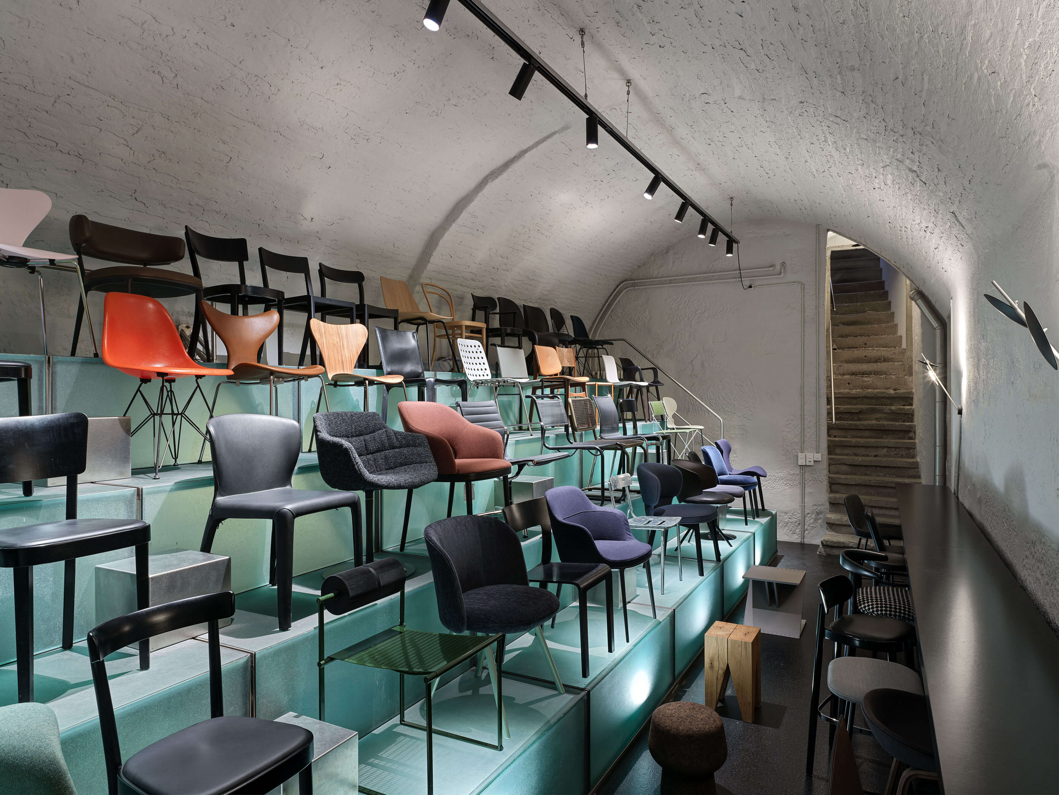 teo jakob Bern Stuhlkeller: Stufenförmige, türkisfarbene Podeste mit vielen unterschiedlichen Designstühlen in verschiedenen Formen, Materialien und Farben unter einer weiss verputzten Gewölbedecke. Rechts verläuft ein schmaler Gang, am Ende führt eine Treppe nach oben.