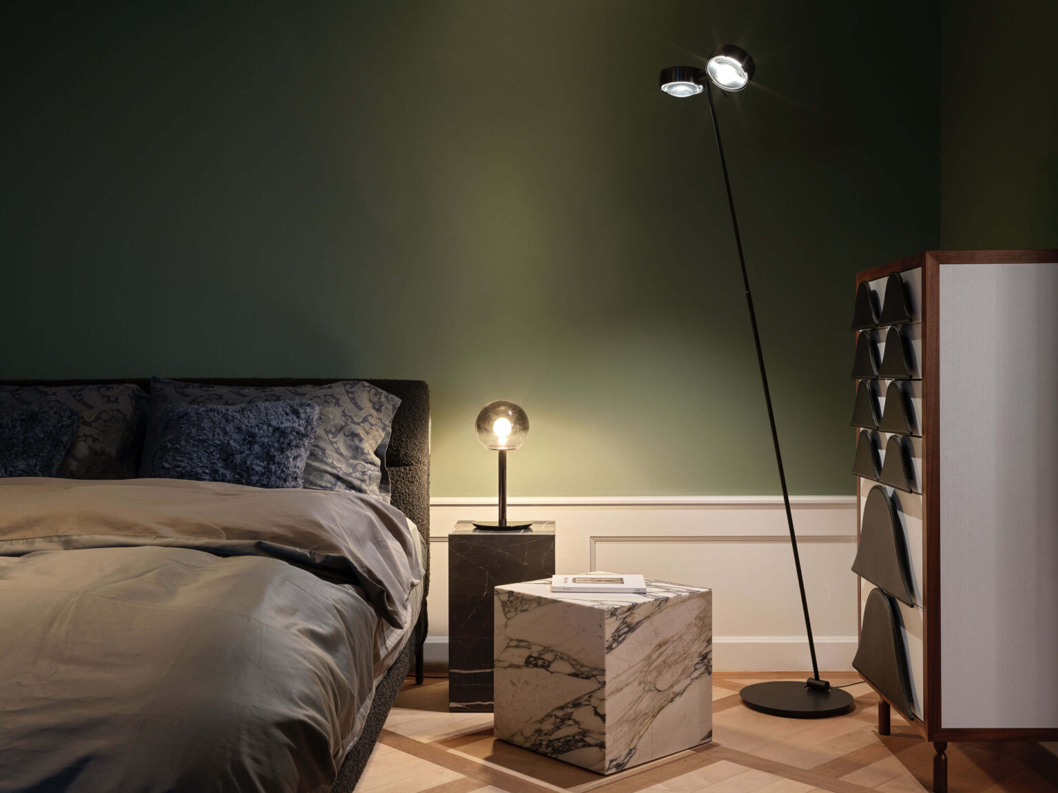 Occhio Light Space, teo jakob Bern, Schlafzimmerinszenierung im Showroom mit Bett, dunkelgrüner Wand, schwarzer Stehleuchte, kleiner Tischleuchte, Marmorsockeln und hellem Sideboard.