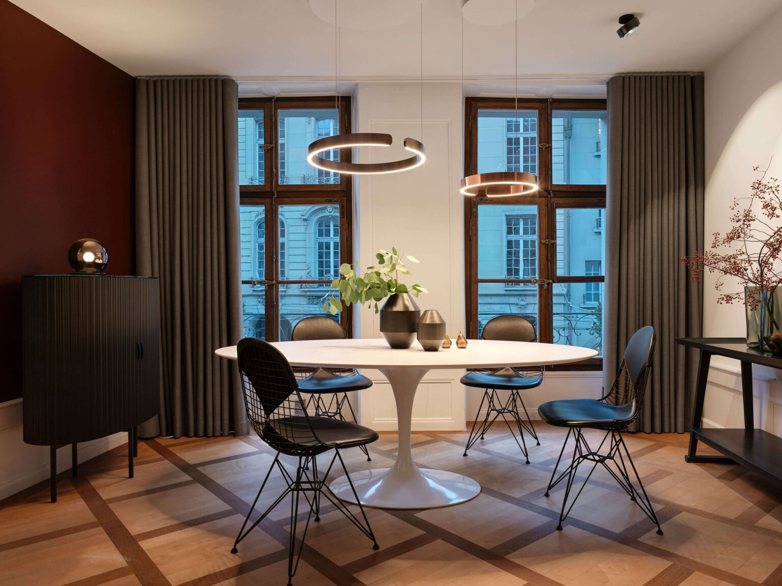 Occhio Light Space bei teo jakob Bern, Essbereich im Showroom mit rundem weissem Tisch, vier schwarzen Stühlen, zwei ringförmigen Pendelleuchten von Occhio, hohen Fenstern und grauen Vorhängen.