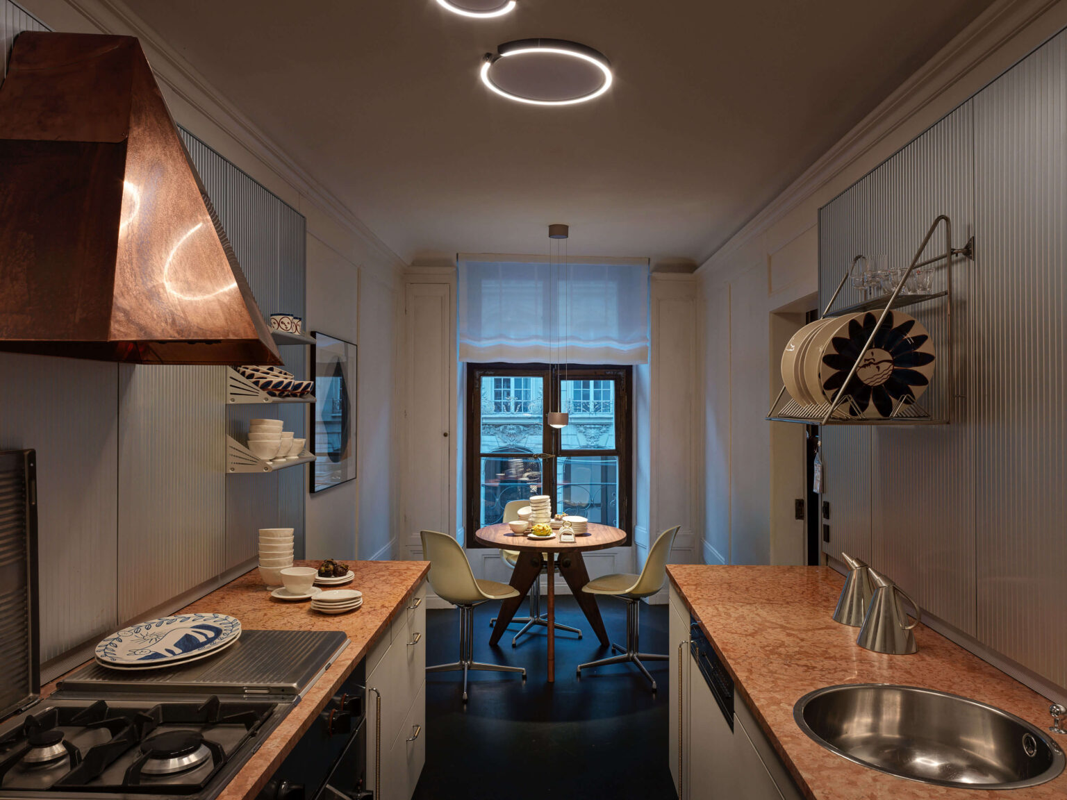 Schmale Küche im Showroom mit zwei Arbeitszeilen, rosafarbenen Arbeitsplatten, rundem Tisch am Fenster und mehreren Leuchten an Decke und über dem Essplatz.