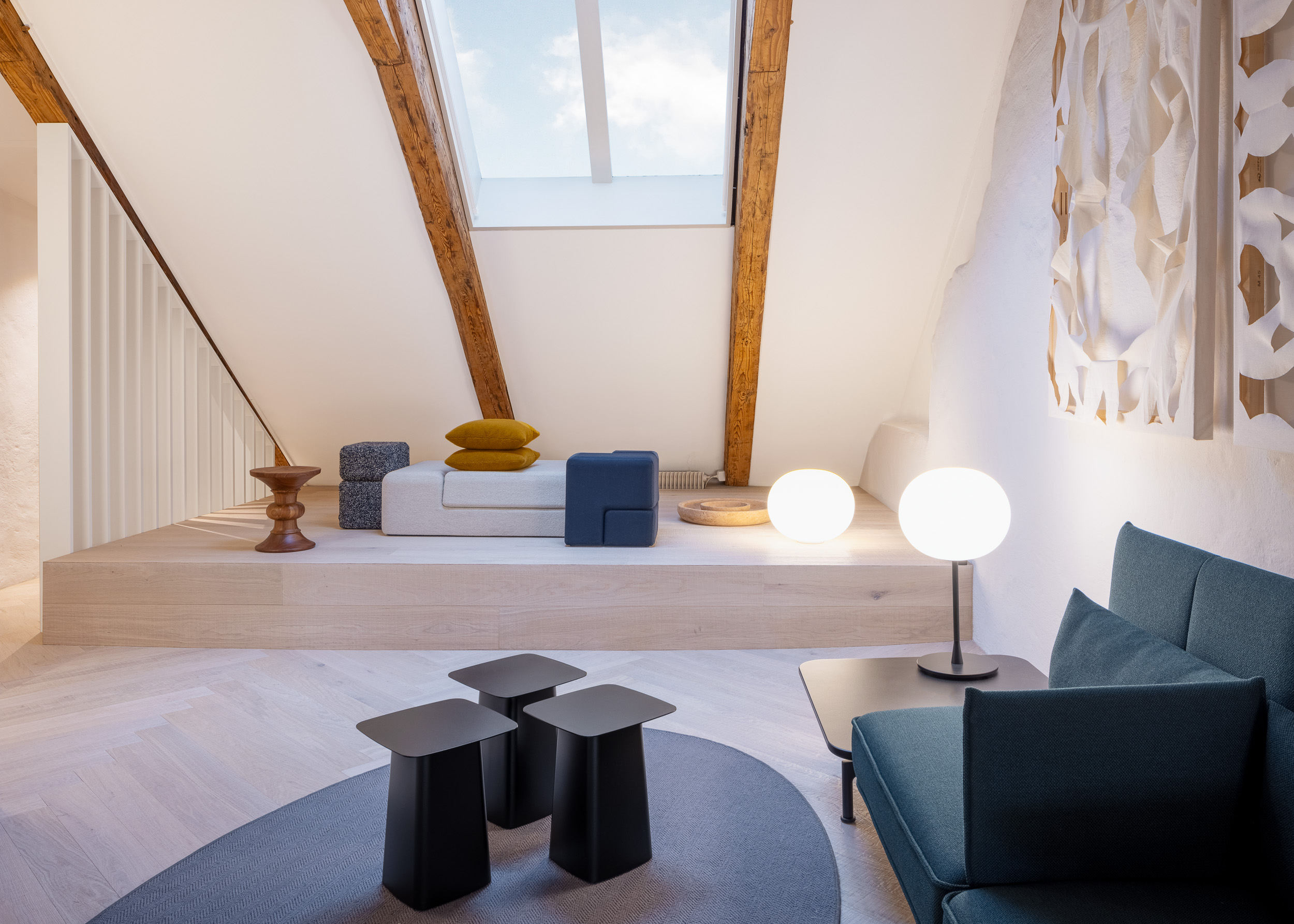 Tageslichtdurchflutetes Dachloft mit sichtbaren Holzbalken: Podest mit Daybed und Kissen, zwei kugelförmige Tischleuchten, vorne drei schwarze Beistelltische.