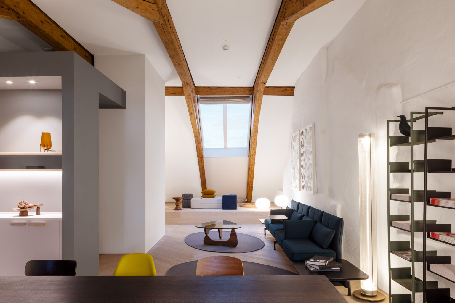 Gesamtblick durchs Loft mit markanten Dachbindern: Podest mit Daybed, runde Teppiche, lange Sofabank, hohe Lichtstele und Regal.