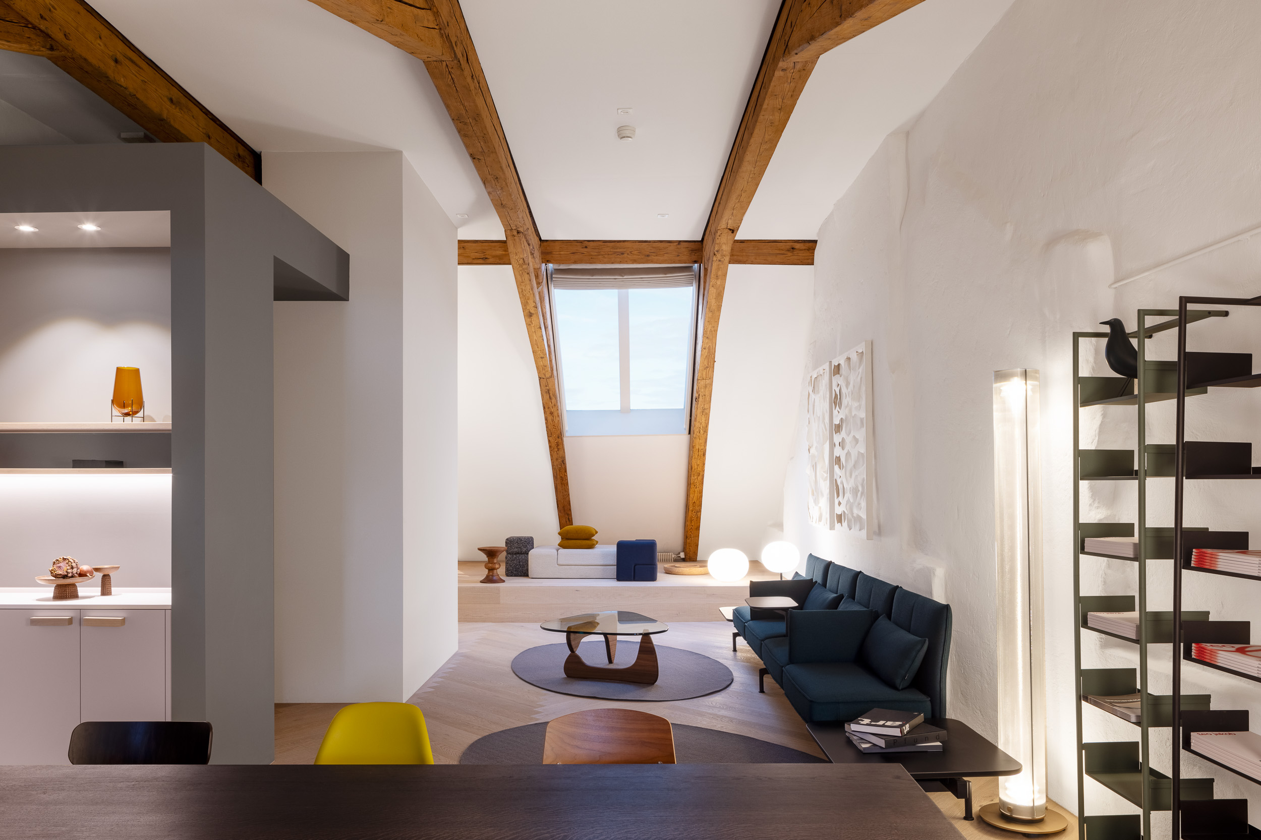 Gesamtblick durchs Loft mit markanten Dachbindern: Podest mit Daybed, runde Teppiche, lange Sofabank, hohe Lichtstele und Regal.