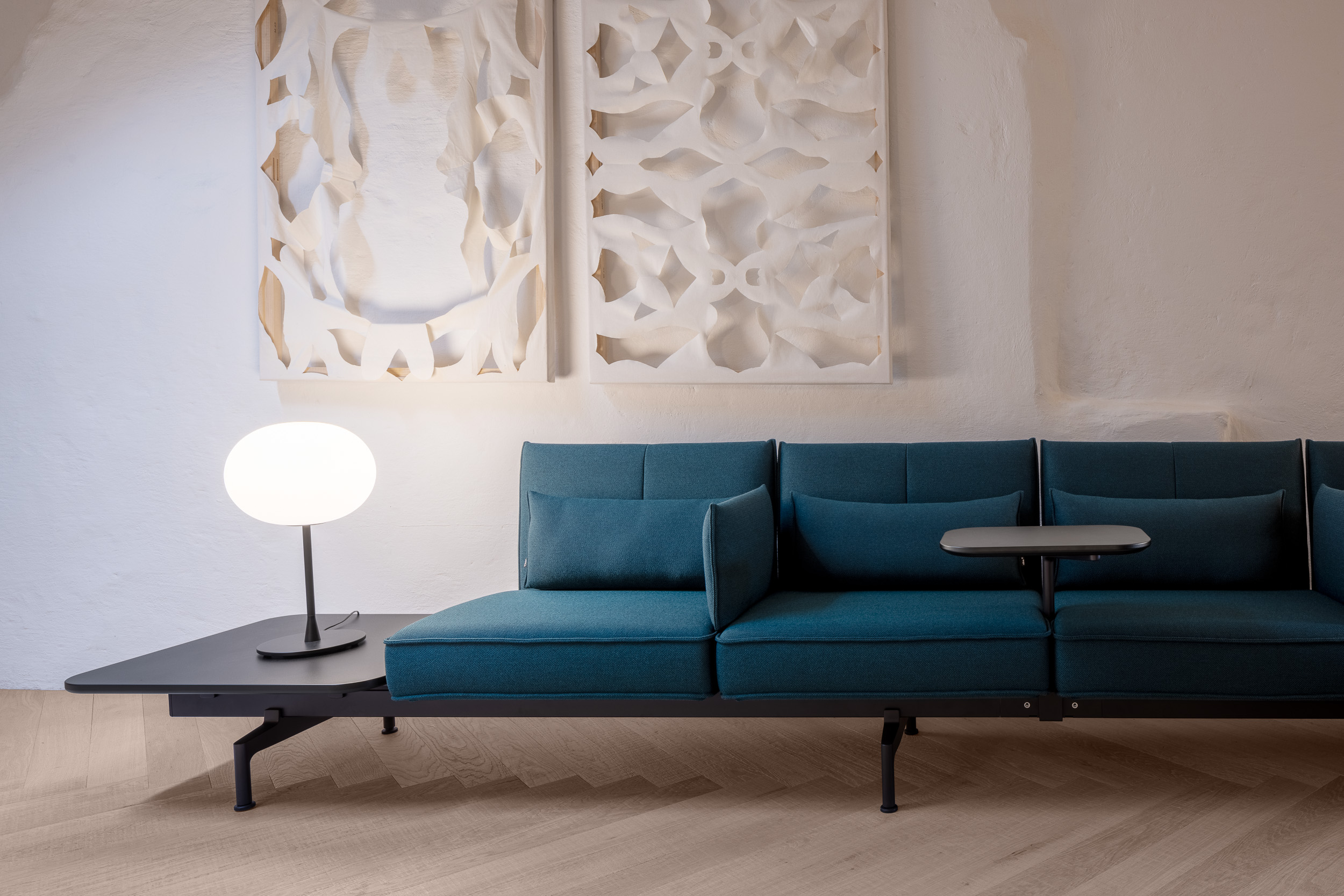 Detail im Loft: petrolfarbene modulare Sofa-Landschaft mit integriertem Ablagetisch, weisser Kugel-Tischleuchte und reliefartigen Wandpaneelen auf hellem Parkett.