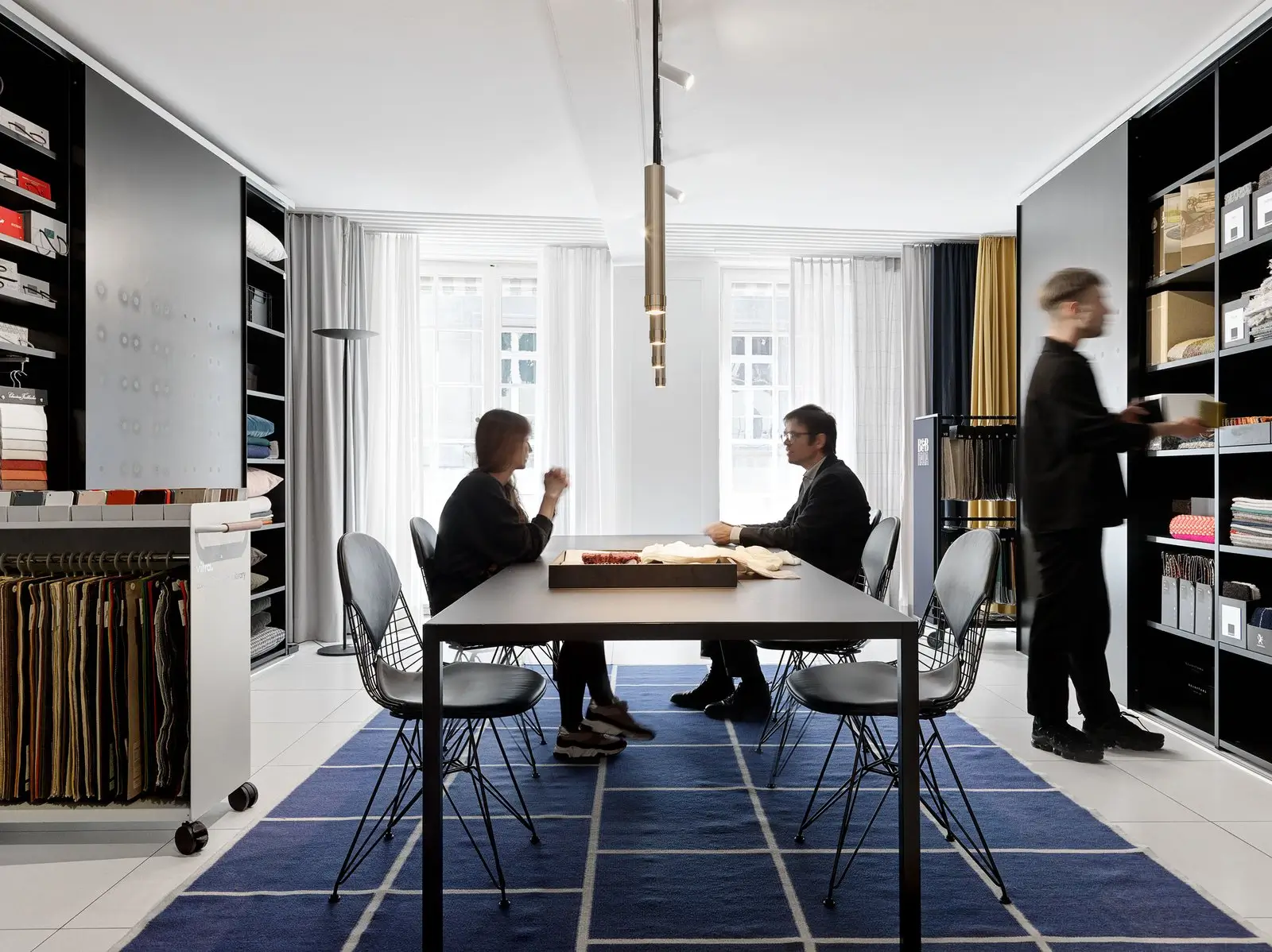 Materialberatung im Showroom: Zwei Personen sitzen an einem langen Tisch auf blauem Teppich mit Raster, umgeben von hohen Regalen mit Stoffmustern und Kollektionsordnern, Pendelleuchte oben und Tageslicht vor grossen Fenstern, eine dritte Person entnimmt Ware aus dem Regal.