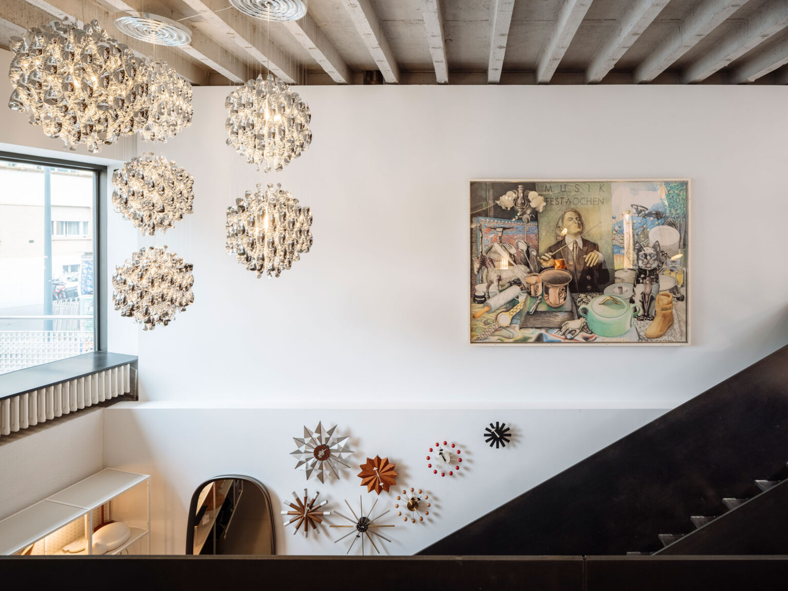 Treppenraum mit mehreren gläsernen Cluster Pendelleuchten, Sichtbetondecke, weisser Wand mit Kunstwerk und Designuhren, Fenster links.
