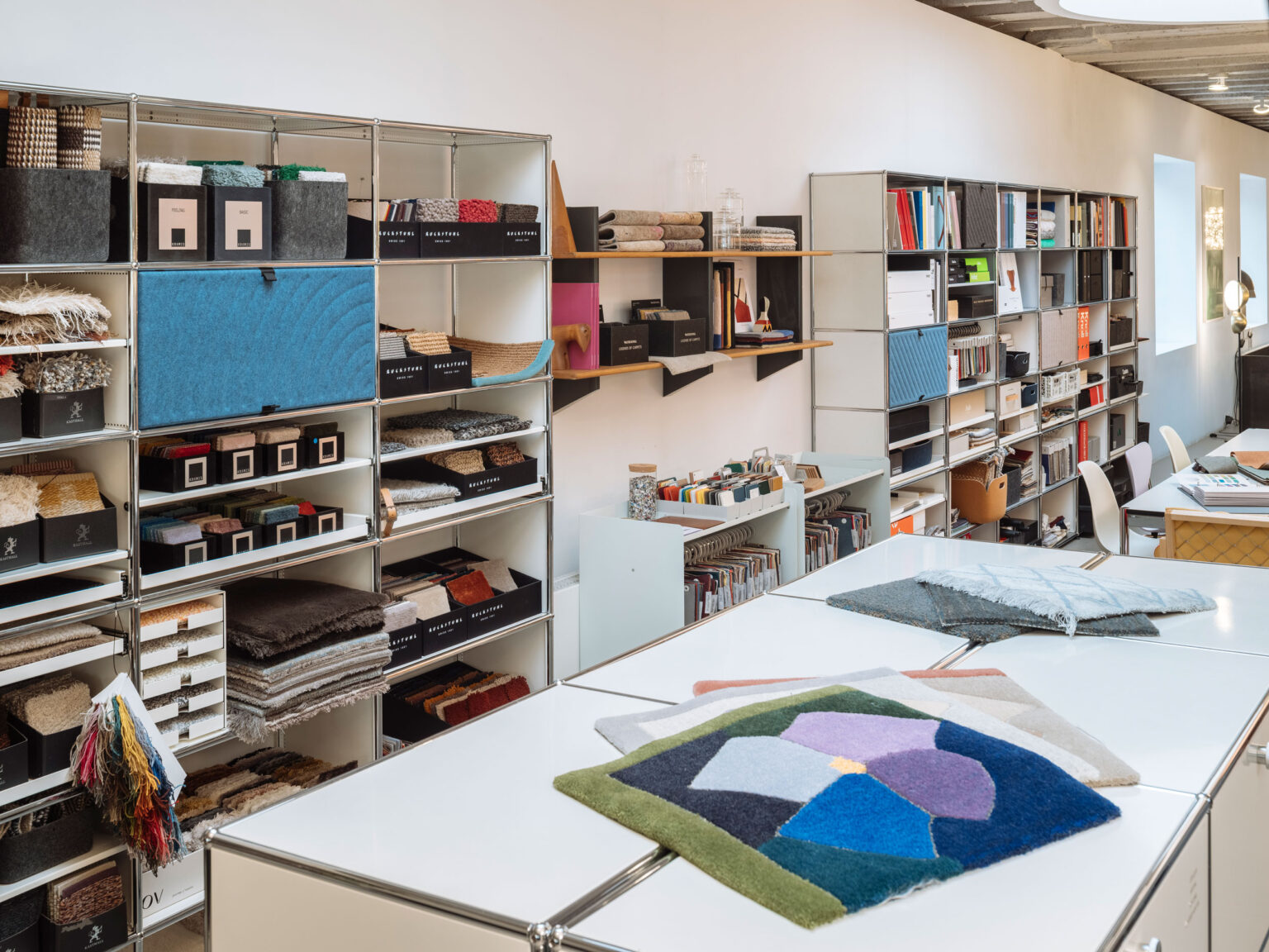 Regalsysteme mit Stoffmustern, Teppichproben und Ordnern, davor grosse Arbeitstische mit farbigen Textil Mustern.