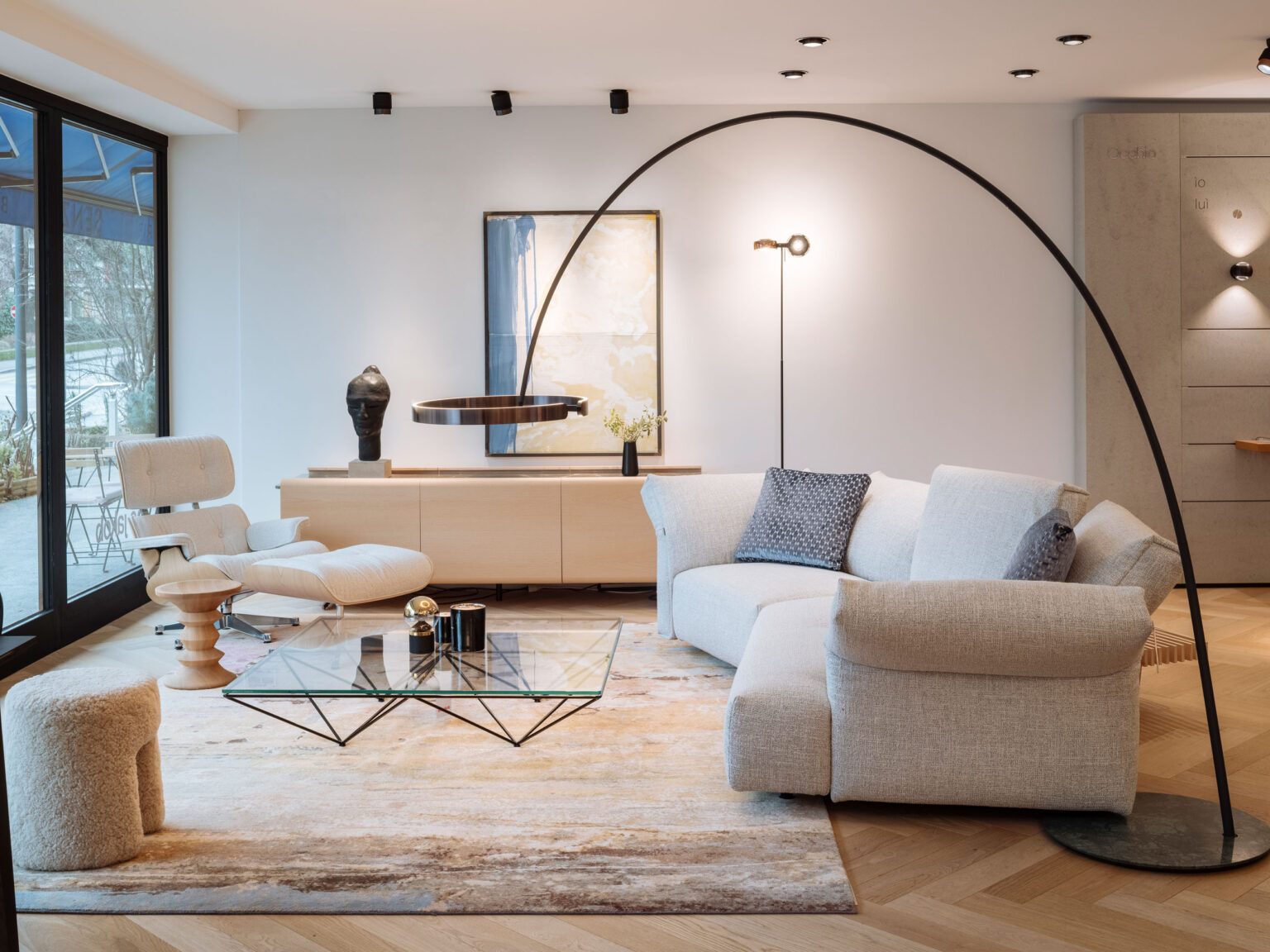 Wohnbereich in der Occhio Gallery im Showroom teo jakob Genf mit hellem Sofa, Lounge Sessel mit Hocker, Glas Couchtisch und grosser bogenförmiger Leuchte über einem hellen Teppich.