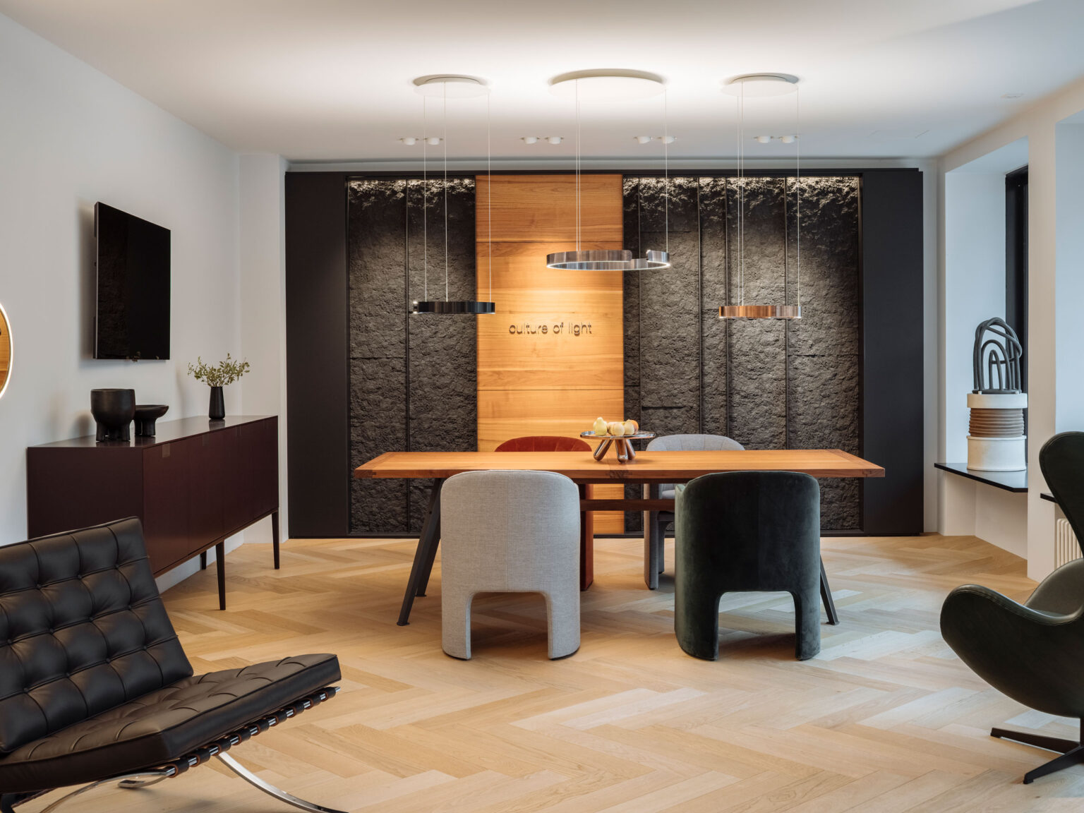 Essbereich in der Occhio Gallery im Showroom teo jakob Genf mit Holztisch, vier gepolsterten Stühlen, drei ringförmigen Pendelleuchten und einer dunklen strukturieren Rückwand mit Holzpaneel.