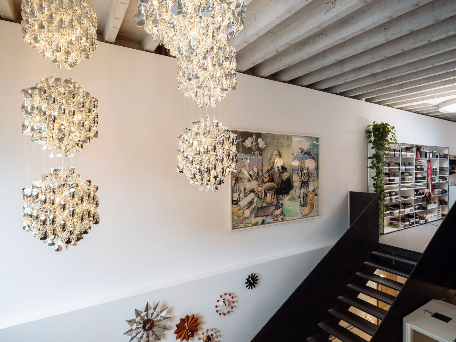 Treppenraum mit mehreren gläsernen Cluster Pendelleuchten, Sichtbetondecke, weisser Wand mit Kunstwerk und Designuhren, Fenster links.