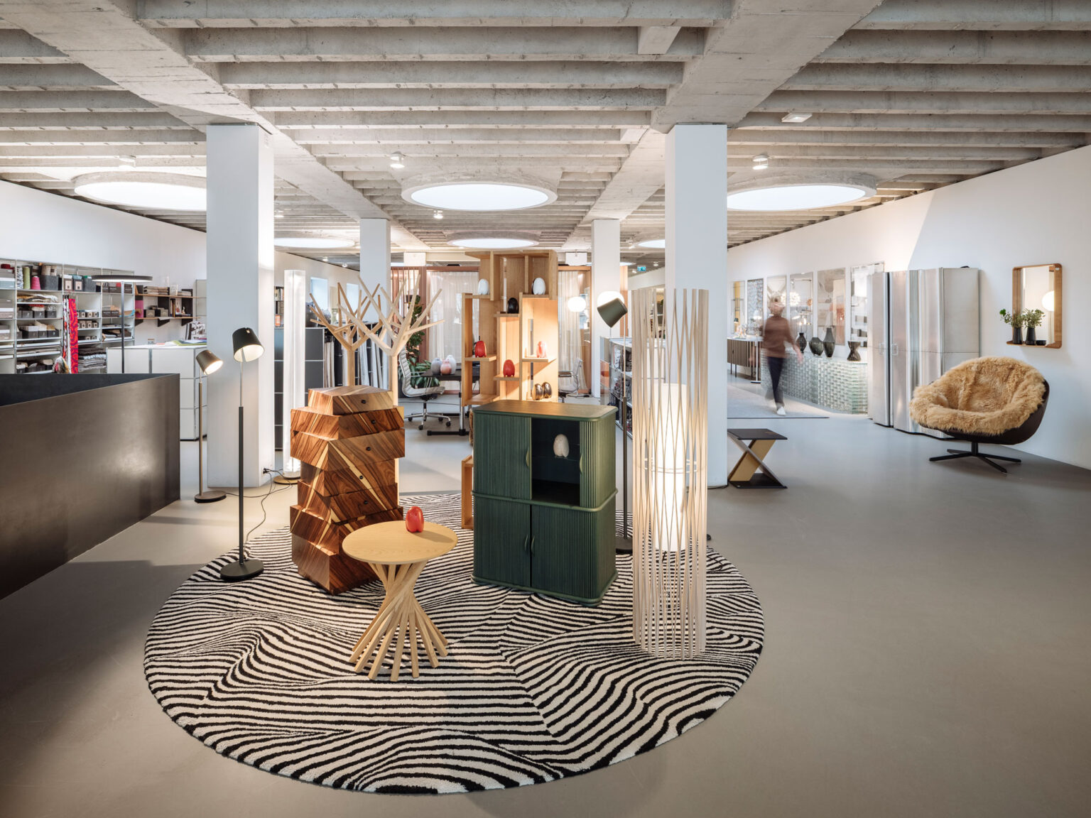 Offene Showroom Fläche mit Holz Garderoben, grünem Sideboard, hoher Stehleuchte, runder gemusterter Teppich und grossen runden Deckenleuchten.