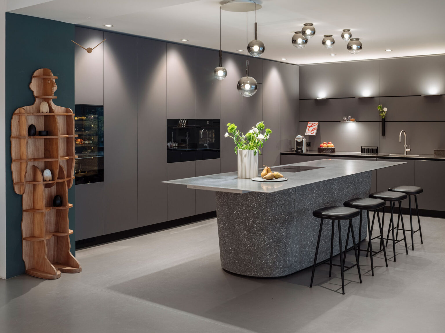 Dunkelgraue Küche mit grosser Steininsel und Barhockern, Hängeleuchten und einer Holzregalfigur an der Wand im Showroom teo jakob Genf.