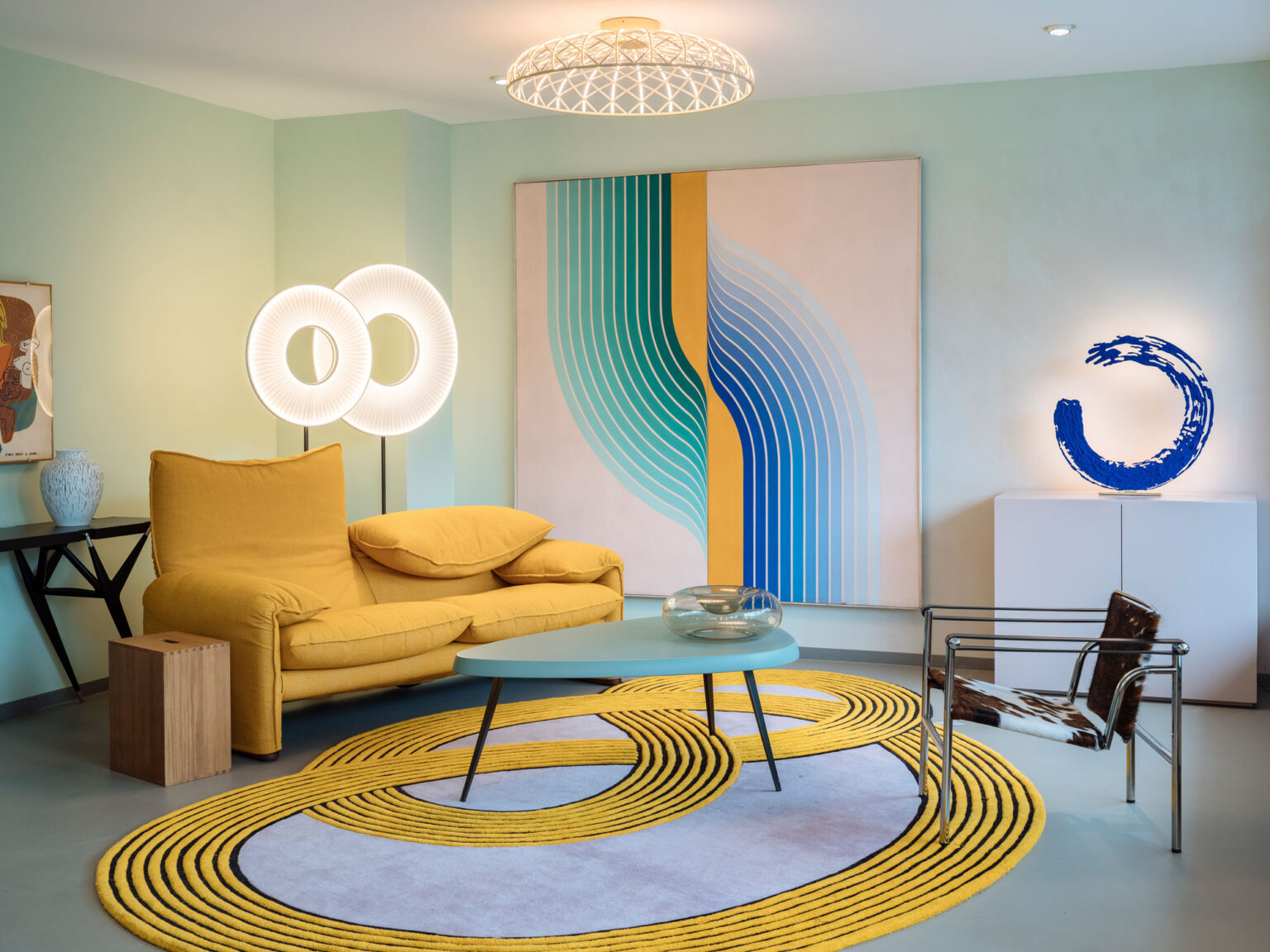 Gelbes Sofa vor grossformatiger Grafik, zwei runde Lichtobjekte, blauer Beistelltisch auf gemustertem Teppich, Kunstobjekt auf Sideboard.