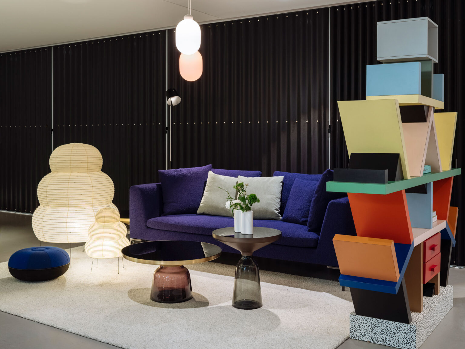 Violette Sofa Lounge vor dunkler Wand mit zwei Papierleuchten, runden Beistelltischen und einer farbigen Regalskulptur im Showroom teo jakob Genf.