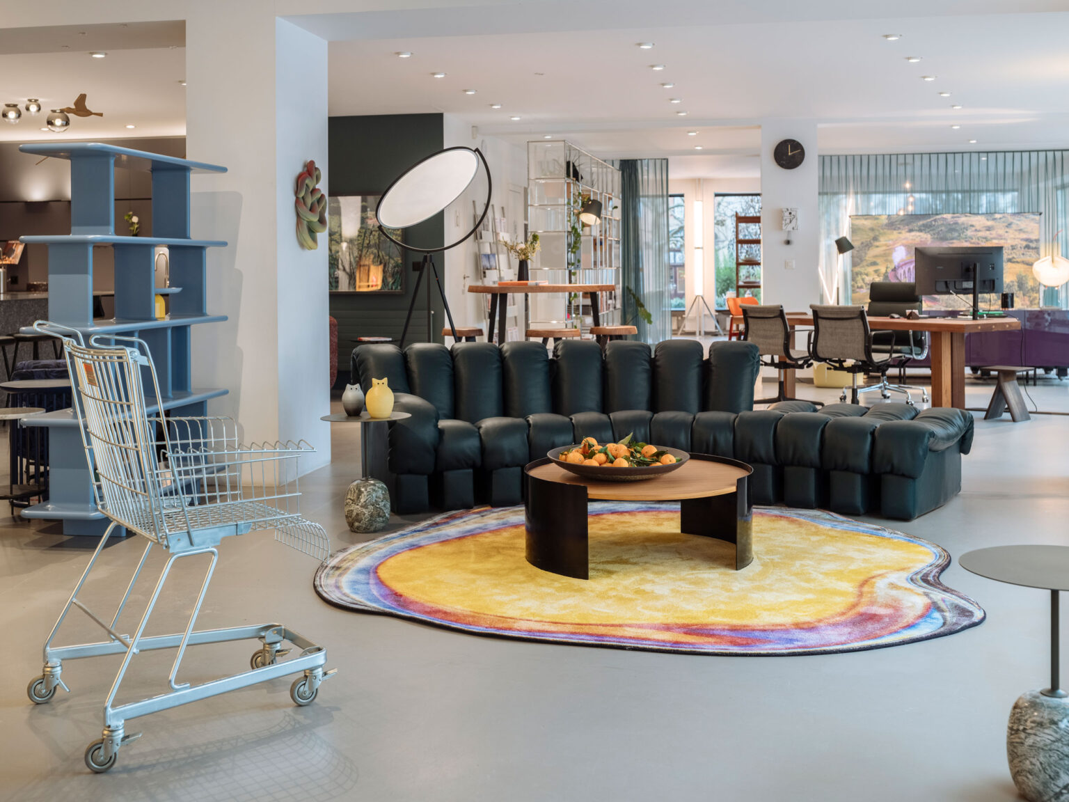 Offener Showroom mit dunkelgrünem Sofa auf buntem Teppich, grosser Studiolampe, Meetingbereich im Hintergrund und Einkaufswagen im Vordergrund.