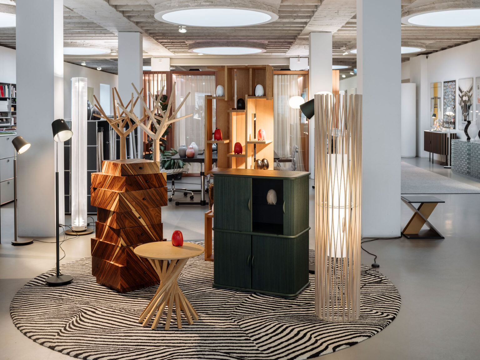 Offene Showroom Fläche mit Holz Garderoben, grünem Sideboard, hoher Stehleuchte, runder gemusterter Teppich und grossen runden Deckenleuchten.