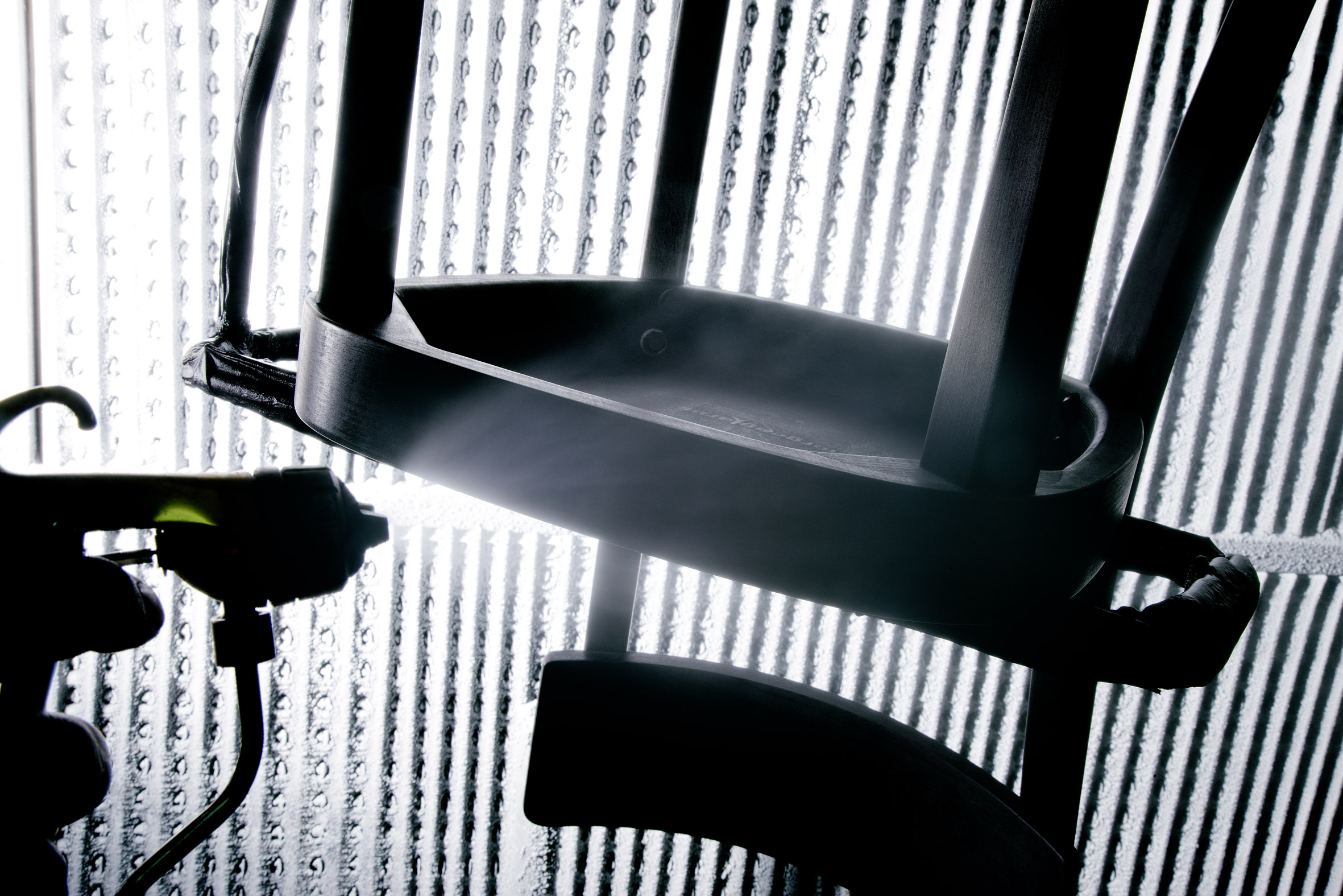 Detailaufnahme eines schwarzen Möbelgestells im Gegenlicht. Im Hintergrund strukturiertes Glas mit vertikalen Rillen. Ein Lichtstrahl betont die Konturen von Sitz und Streben. Links sind weitere dunkle Elemente nur schemenhaft sichtbar.