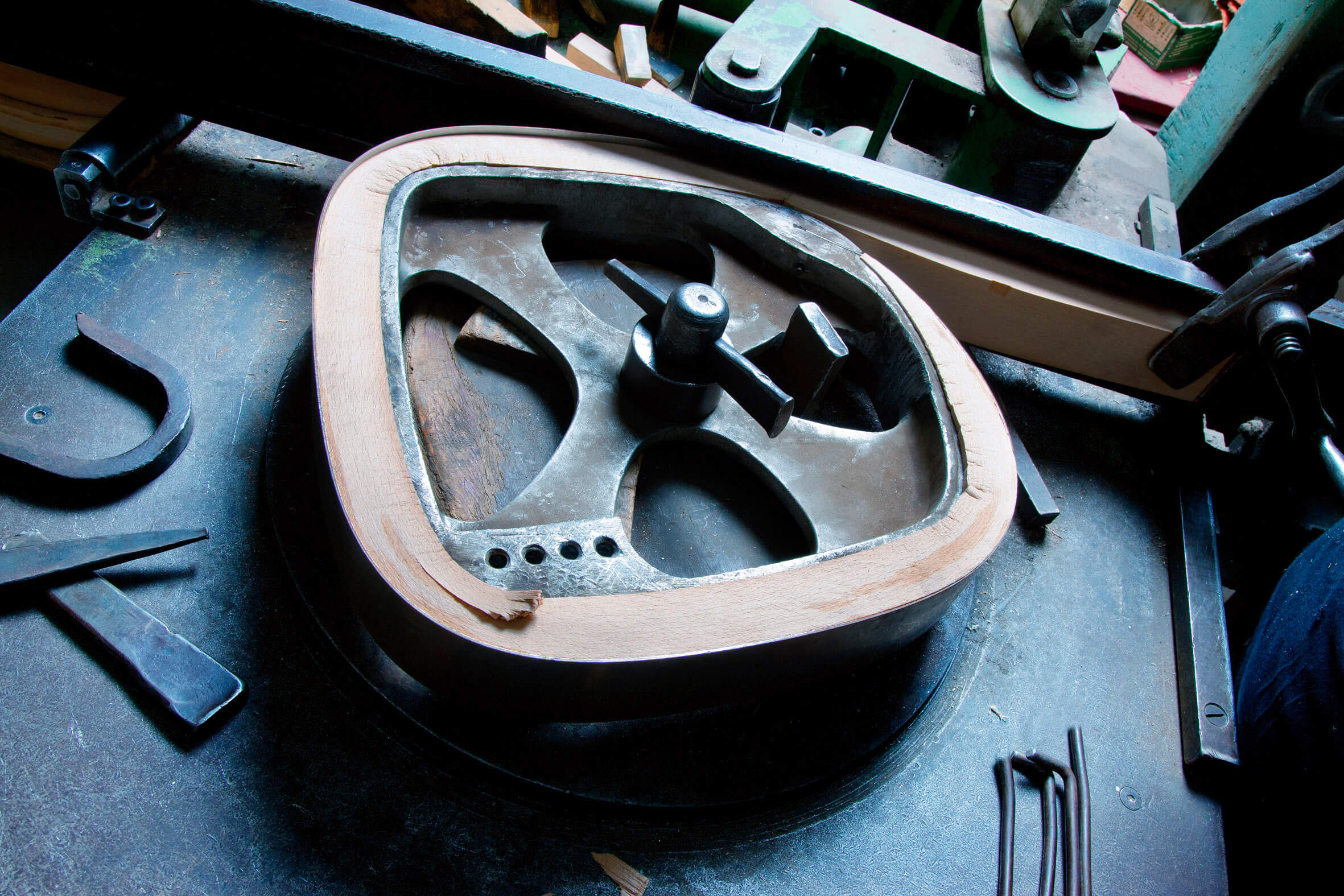 Werkstattaufnahme einer Metallform mit eingespanntem, gebogenem Holzrahmen. Die Form liegt auf einer Maschine oder Werkbank, daneben sind Werkzeuge und Metallteile verteilt. Der Holzrahmen zeigt eine glatte, helle Oberfläche mit sichtbarer Kante und kleiner Ausbruchstelle.