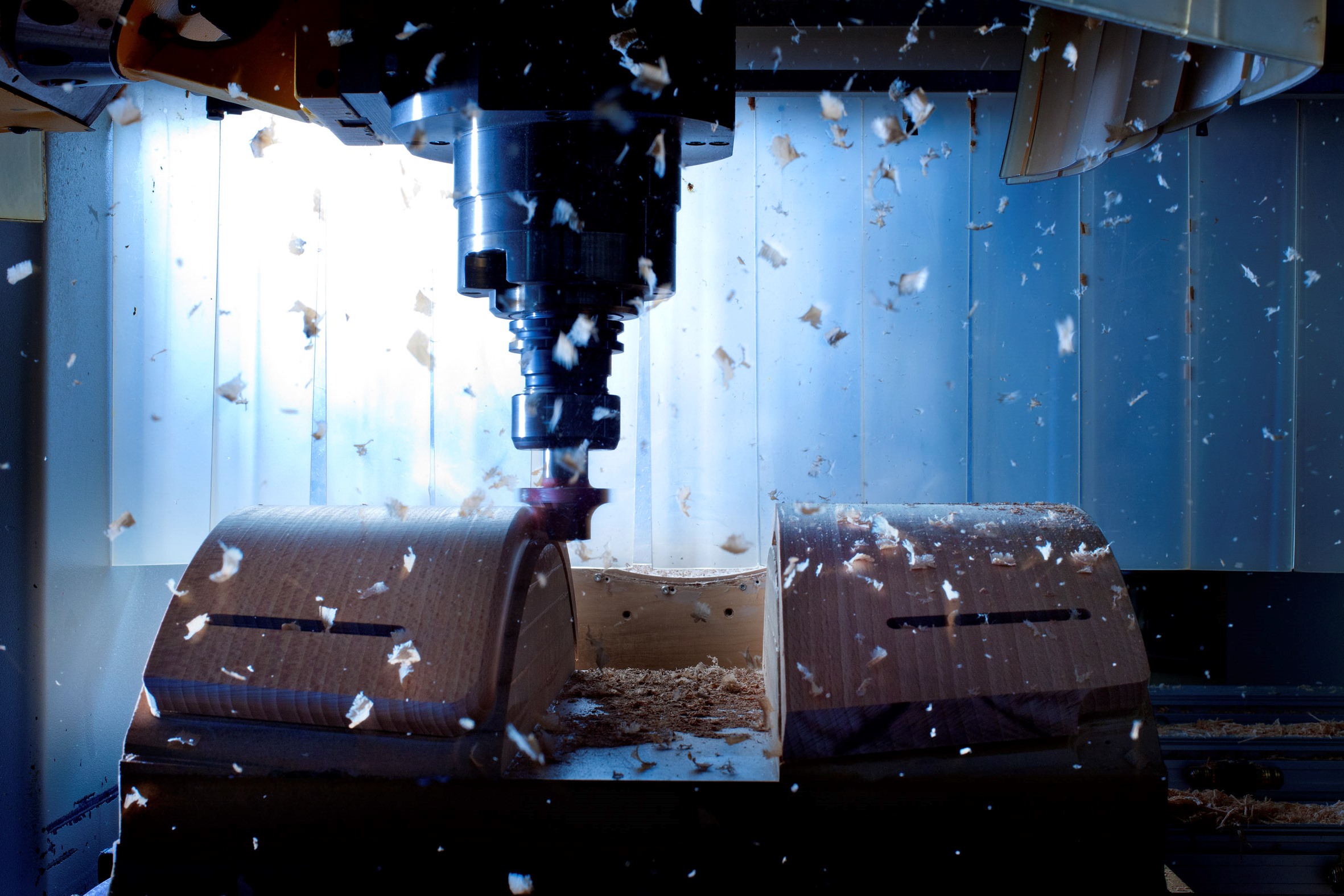 CNC Fräse in Betrieb, die ein Holzteil bearbeitet. Der Fräskopf steht mittig über dem Werkstück, Holzspäne fliegen durch den Innenraum der Maschine. Zwei geformte Holzblöcke liegen links und rechts auf dem Arbeitstisch. Blaues Maschinengehäuse und Spänesammlung am Boden sind sichtbar.