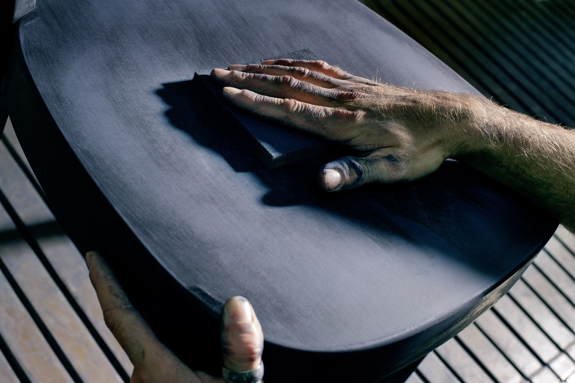 Nahaufnahme einer Hand, die eine dunkle Holzfläche mit Schleifpapier bearbeitet. Die geschwungene Form wirkt wie eine Sitzschale. Eine zweite Hand stabilisiert das Werkstück am Rand. Licht und Schatten betonen die feine Maserung und den Schleifprozess.