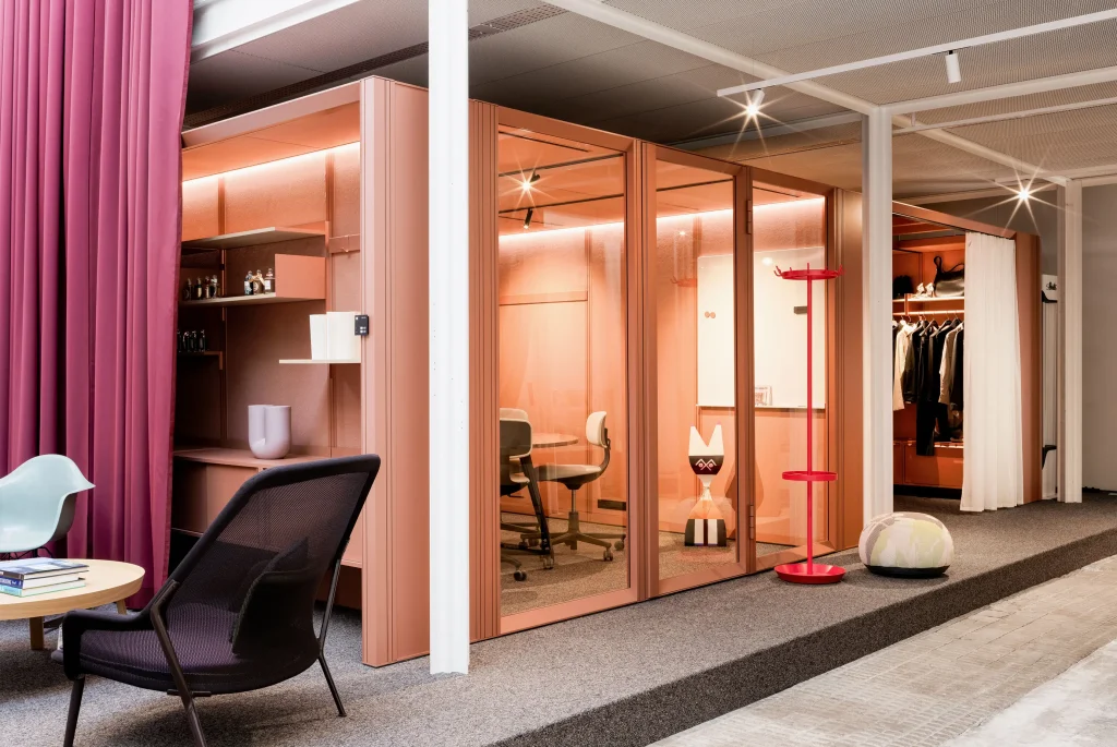 Innenansicht mit terrakottafarbenen Meetingboxen mit Glasfront, integrierten Regalen und Whiteboard, davor Loungeecke mit schwarzem Netzsessel und runder Tisch, links bordeauxfarbener Vorhang, rechts roter Garderobenständer und offene Garderobe und warme Beleuchtung.