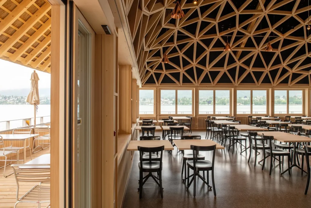 Restaurantbereich mit polygonaler Holzdecke, Reihen heller Tische und schwarzer Stühle, grosse Fenster mit Blick auf den See, links vorgelagerte Terrasse mit Metallmöbeln.