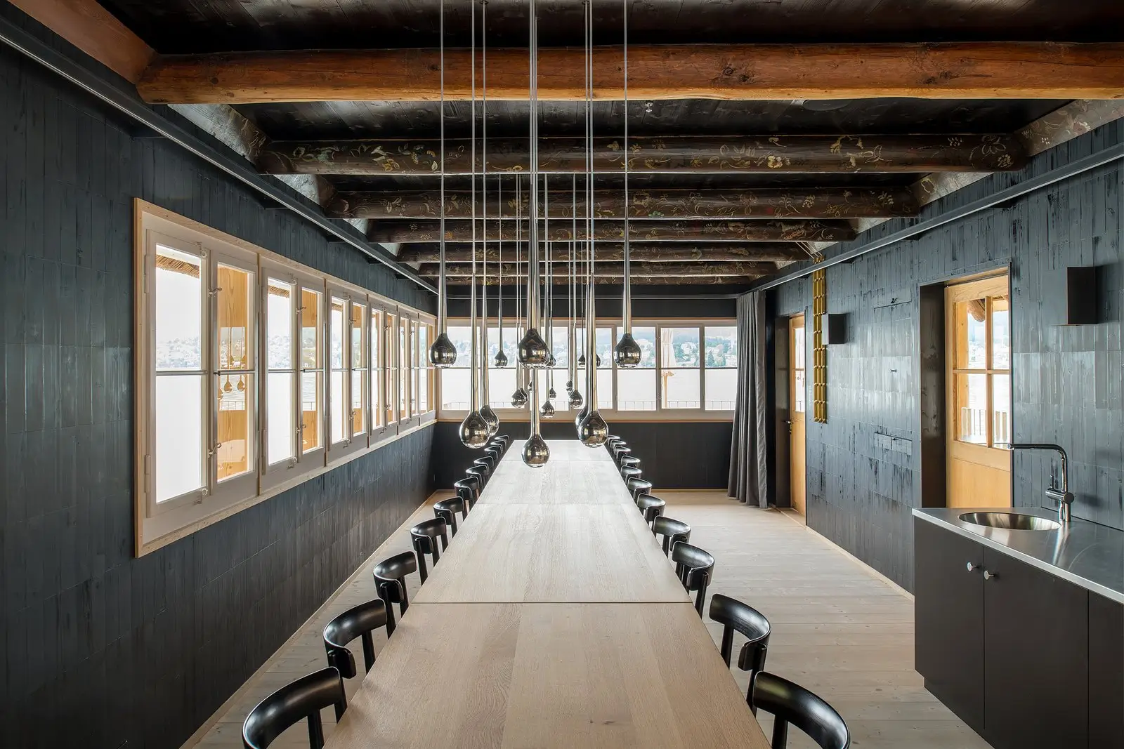 Langer Besprechungstisch mit schwarzen Stühlen in holzverkleidetem Raum, zahlreiche tropfenförmige Pendelleuchten hängen in Reihen von der Balkendecke, seitlich Fensterfront mit Seeblick, rechts kleine Küche mit Spüle.
