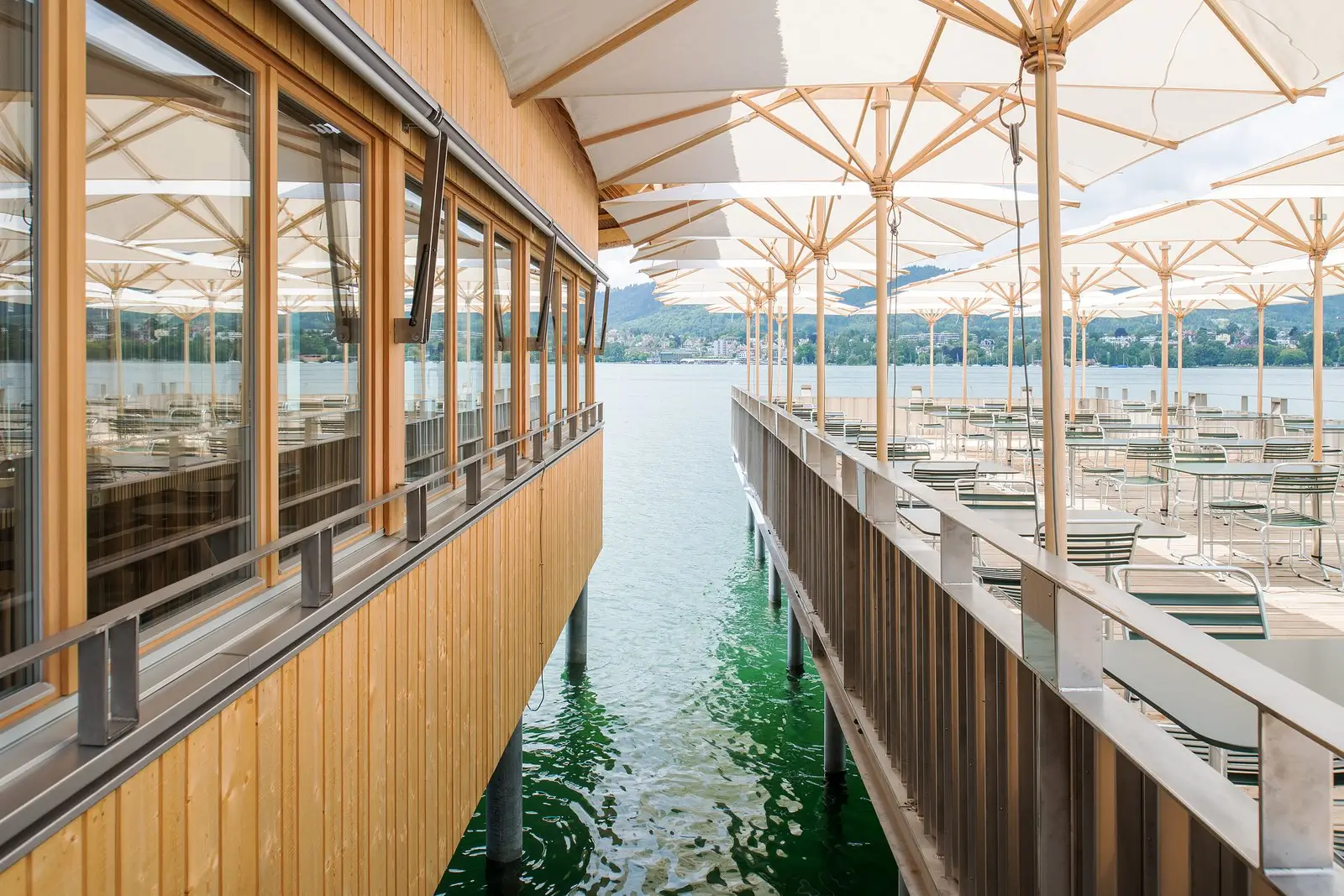 Holzbau auf Stelzen am See mit seitlicher Fensterfront und angrenzender Sonnenterrasse unter weissen Schirmen, Reihen aus Metalltischen und -stühlen über grünem Wasser mit Blick auf Ufer und Hügel.