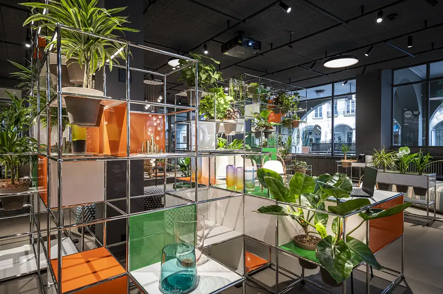 Showroombereich von teo jakob mit modularen Metallregalen in Chrom, bestückt mit vielen Zimmerpflanzen, Vasen und grüne sowie orange Fachmodule; dahinter grosse Fenster mit Laubengang, dunkle Decke mit Spotbeleuchtung.