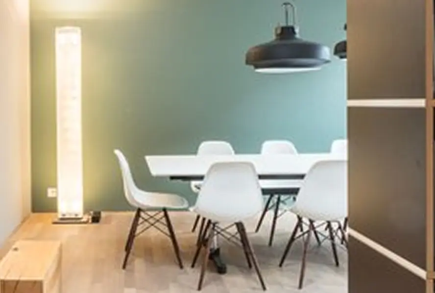 Moderner Essbereich mit weissem Tisch und sechs weissen Schalenstühlen auf Holzgestell, zwei schwarzen Pendelleuchten darüber, links eine hohe Lichtsäule vor grün gestrichener Wand, heller Holzboden.