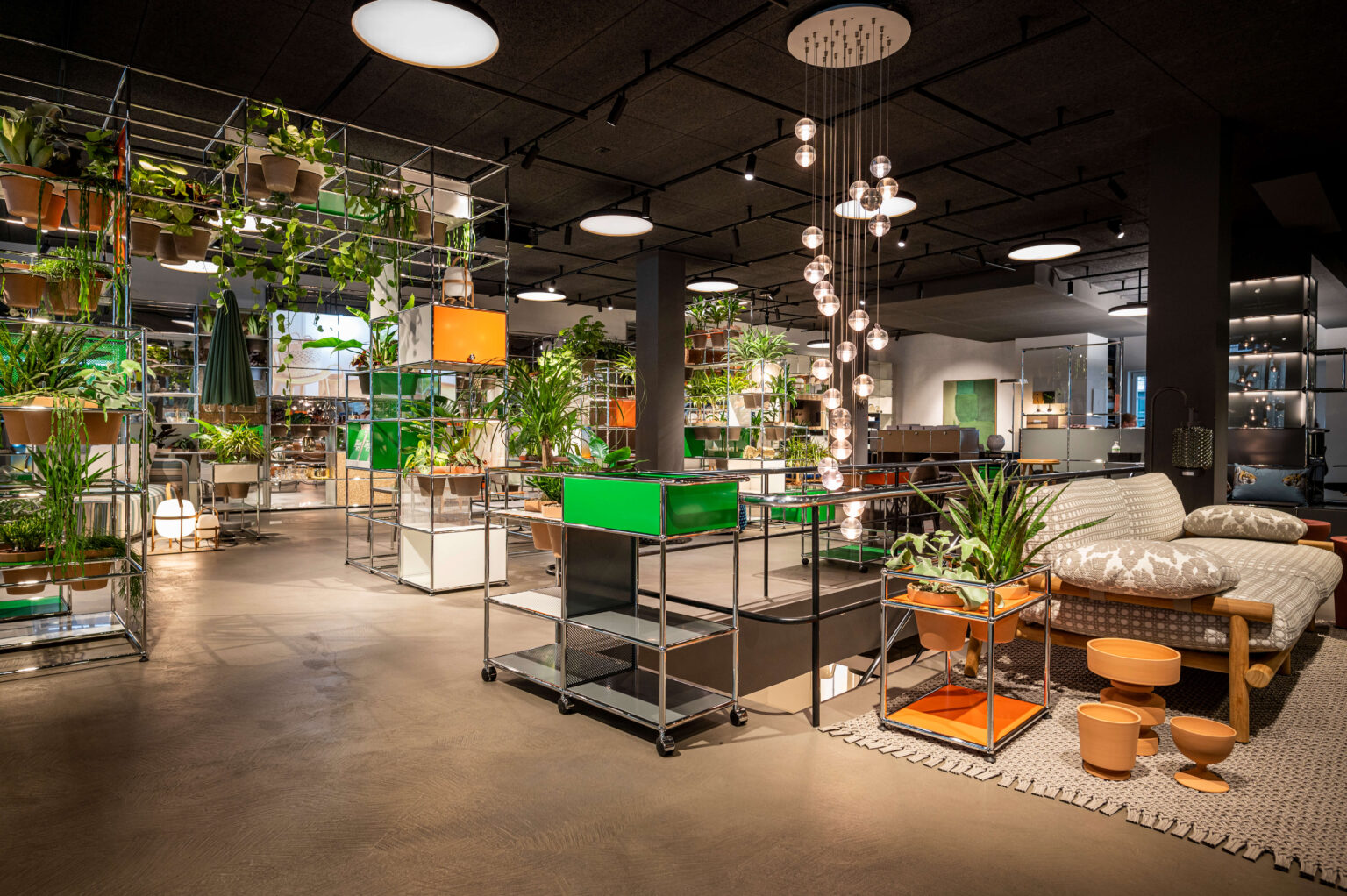 Grosszügiger Showroom mit modularen Metallregalen voller Grünpflanzen, hängenden Glaskugelleuchten, Rollwagen mit grünen und orangefarbenen Elementen sowie Sofa und terrakottafarbenen Töpfen im Vordergrund.