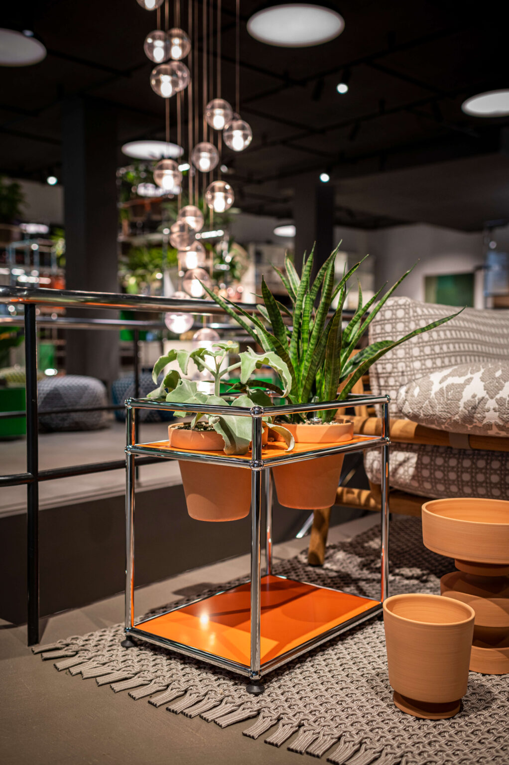 Chromfarbener Rollwagen mit orangefarbenen Ablagen und terrakottafarbenen Töpfen mit Grünpflanzen, inszeniert im Showroom vor hängenden Glaskugelleuchten, Teppich und gemusterter Sitzbank.