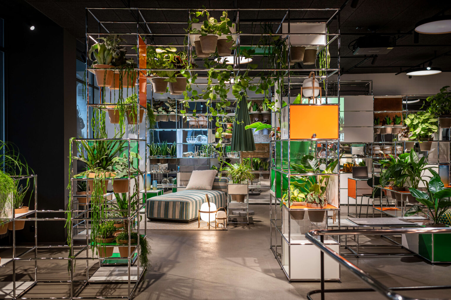 Showroom mit modularen Metallregalen und vielen grünen Pflanzen. In der Mitte steht eine Liege mit gestreiftem Bezug, umgeben von Accessoires und orangefarbenen Elementen in den Regalsystemen.