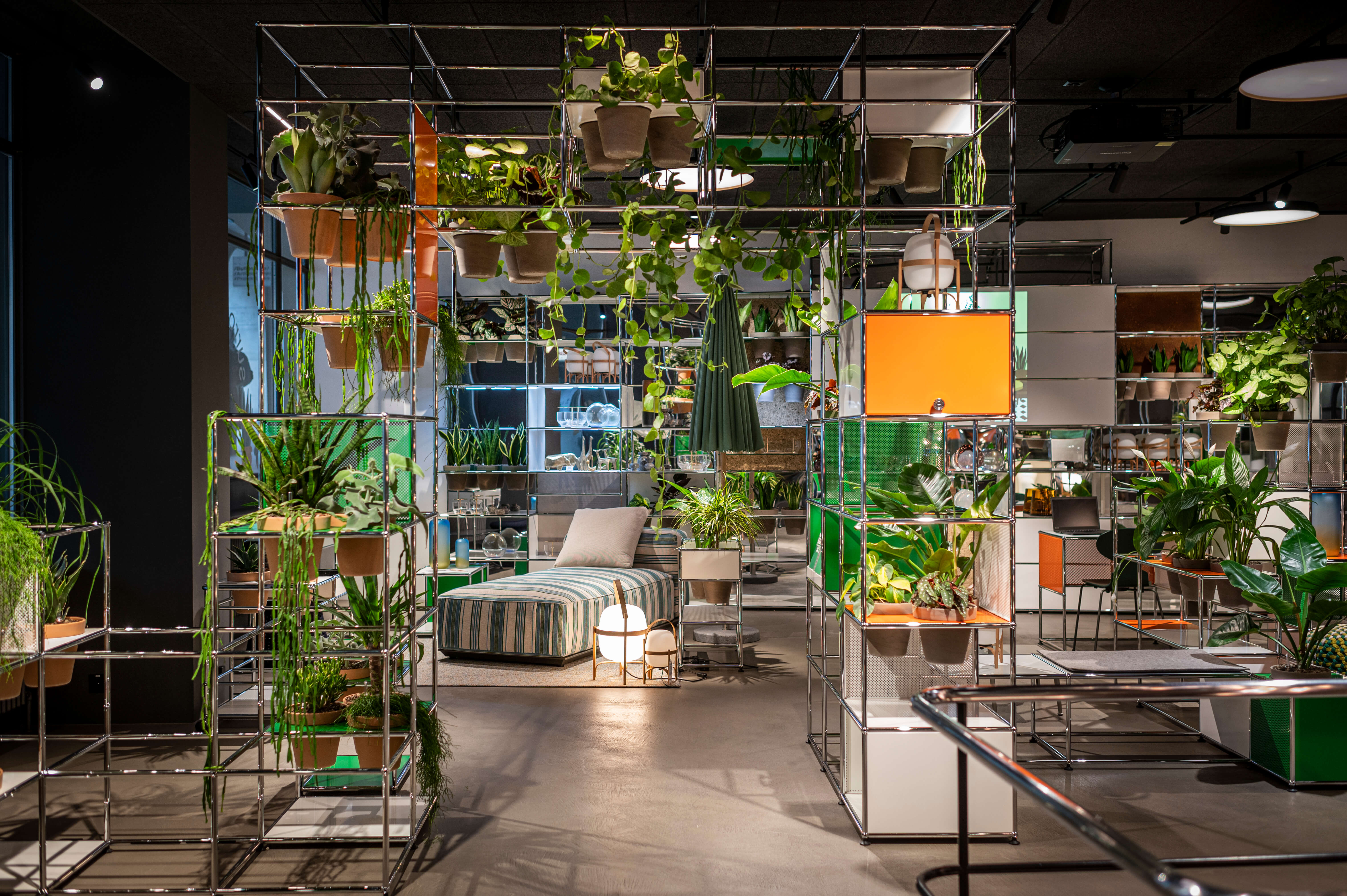 Showroom mit modularen Metallregalen und vielen grünen Pflanzen. In der Mitte steht eine Liege mit gestreiftem Bezug, umgeben von Accessoires und orangefarbenen Elementen in den Regalsystemen.