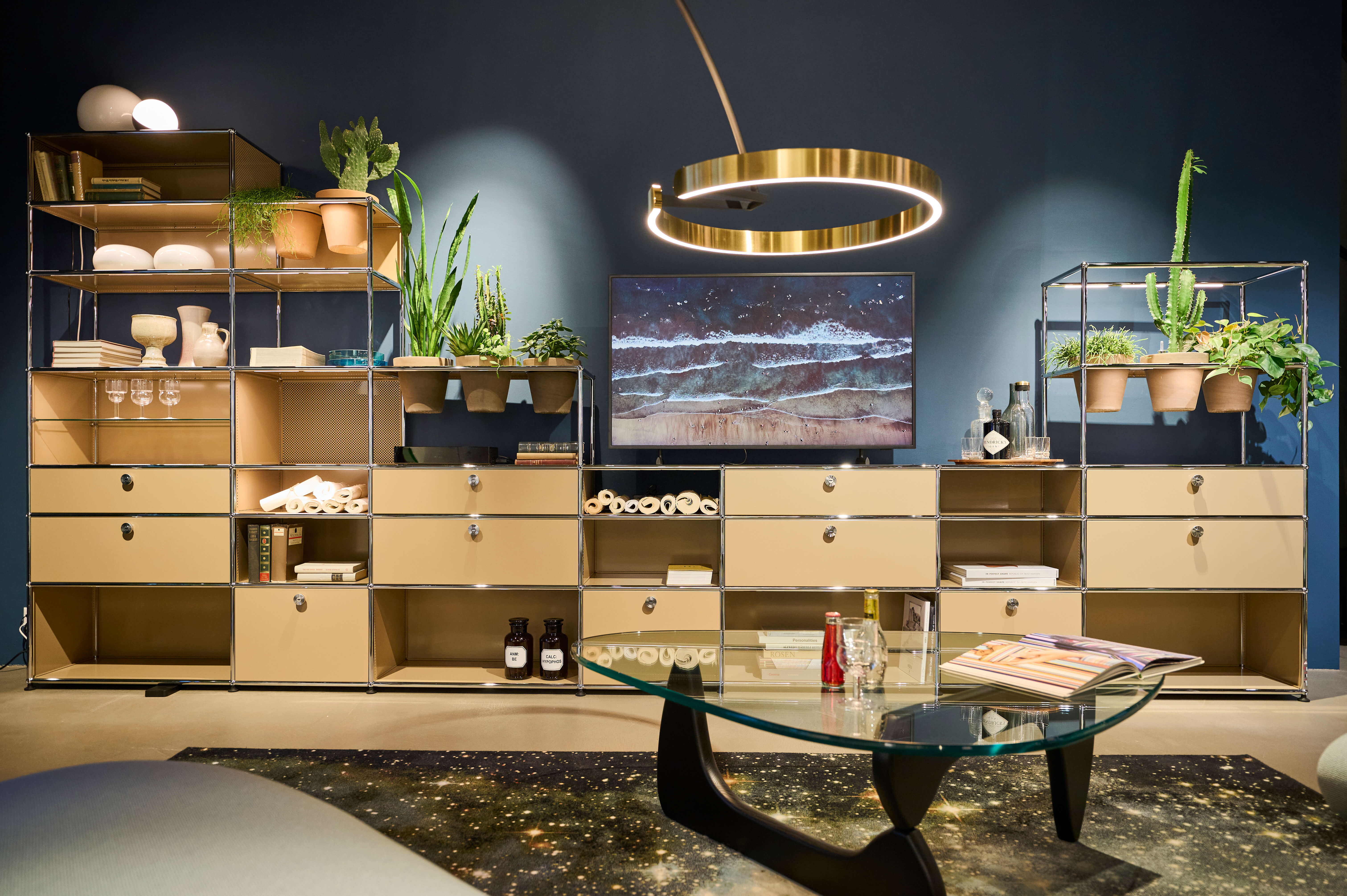 Showroom Szene mit modularen Metallregalen in Beige, dekoriert mit Pflanzen, Geschirr und Accessoires. Zentral hängt ein TV vor dunkelblauer Wand, darüber eine runde Messing Pendelleuchte. Im Vordergrund steht ein runder Glastisch mit Zeitschriften und Flaschen auf einem dunklen Teppich.