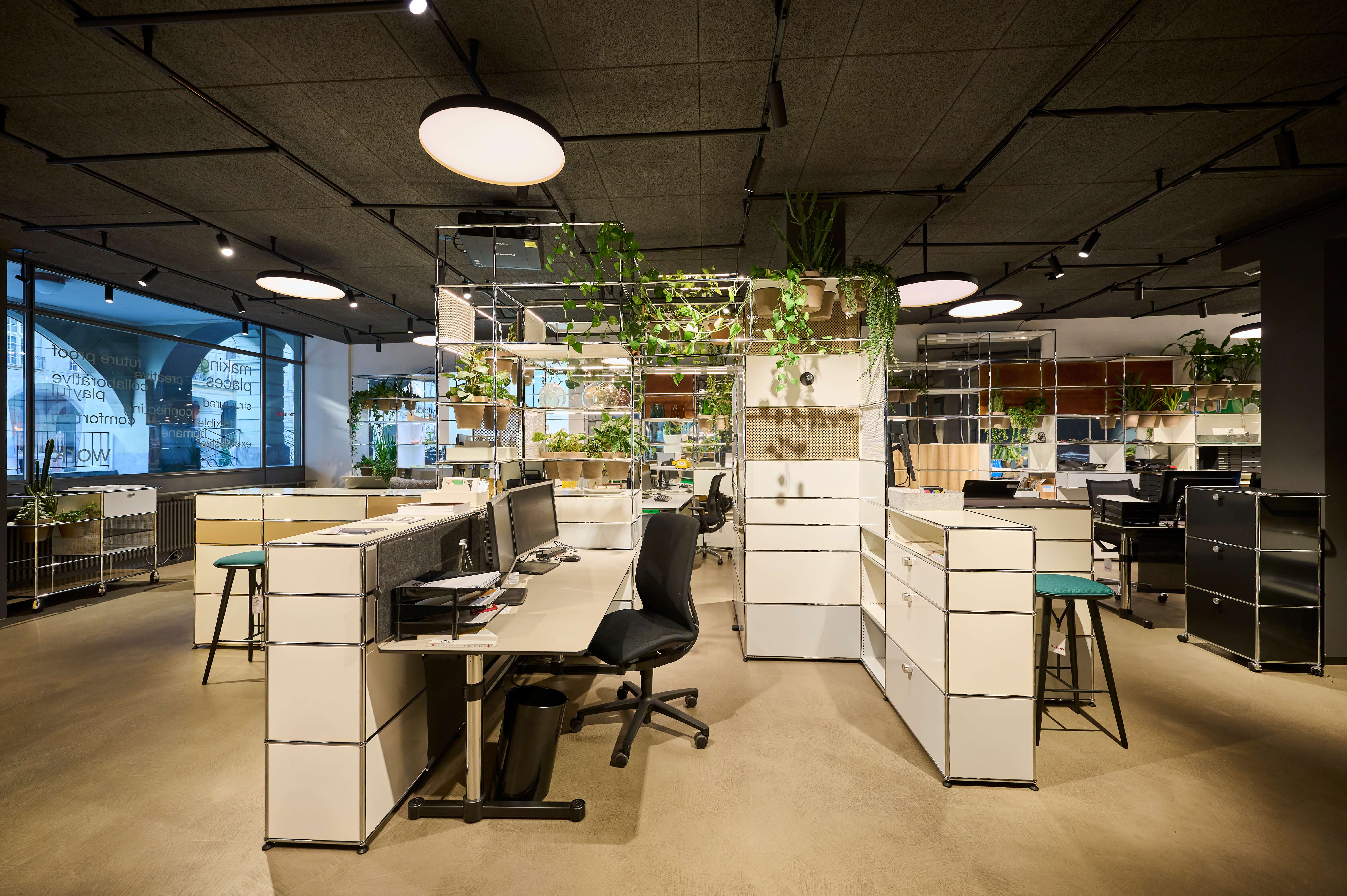 Offener Buero Bereich mit modularen weissen Sideboards und Arbeitsplätzen. Mehrere Bildschirme und Drehstühle stehen zwischen den Möbelinseln. Hängepflanzen und Regale strukturieren den Raum. Runde Deckenleuchten und Schienenstrahler sorgen für gleichmässiges Licht. Links sorgt eine grosse Fensterfront für Tageslicht.