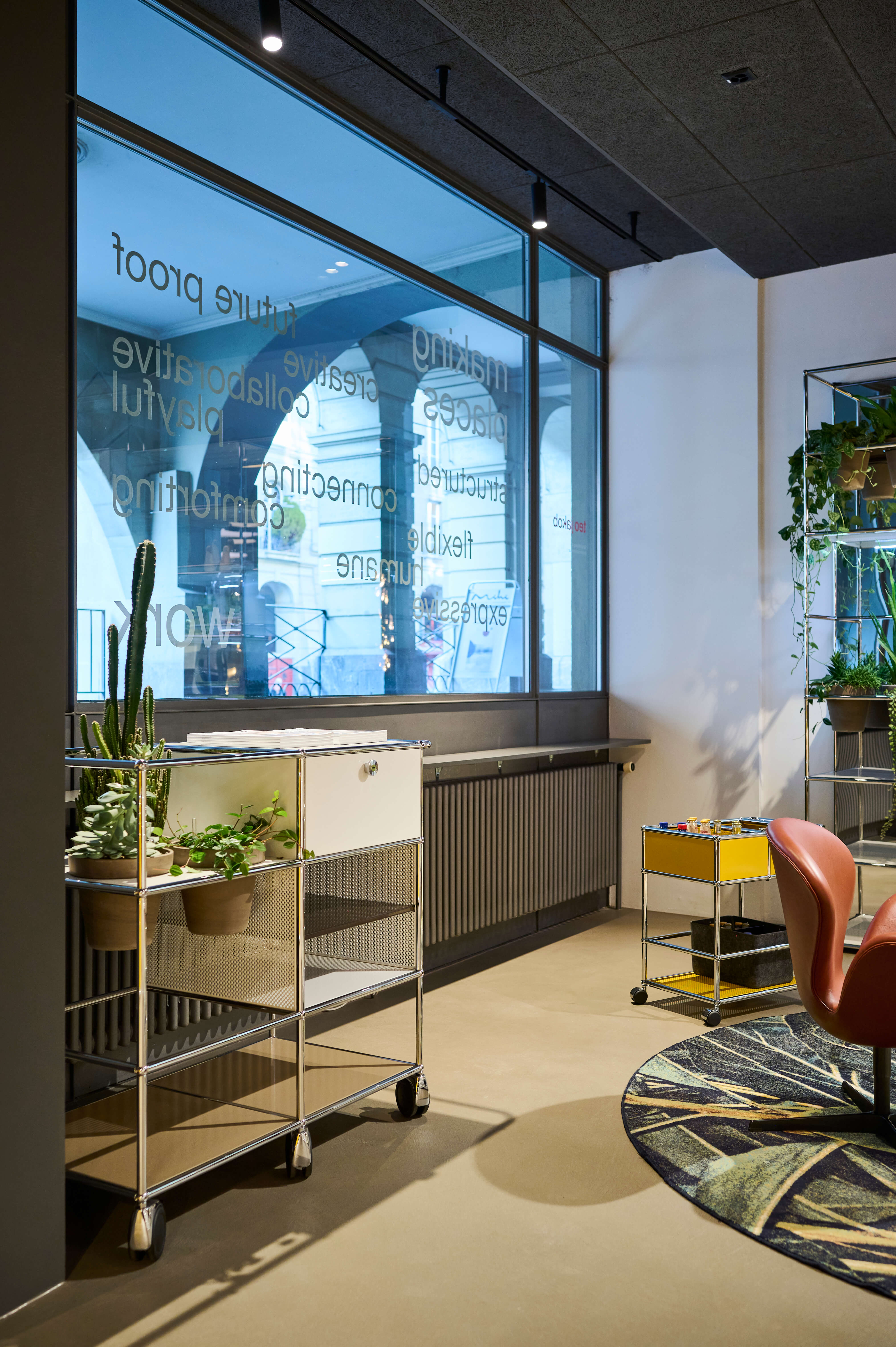 Moderner Showroom Bereich mit grossem Fenster und typografischer Folienbeschriftung, davor zwei rollbare Servierwagen mit Pflanzen, gelbem Einsatz und Ablagefächern. Im Hintergrund stehen ein Ledersessel, ein runder Teppich und eine hohe Pflanze unter Schienenbeleuchtung.