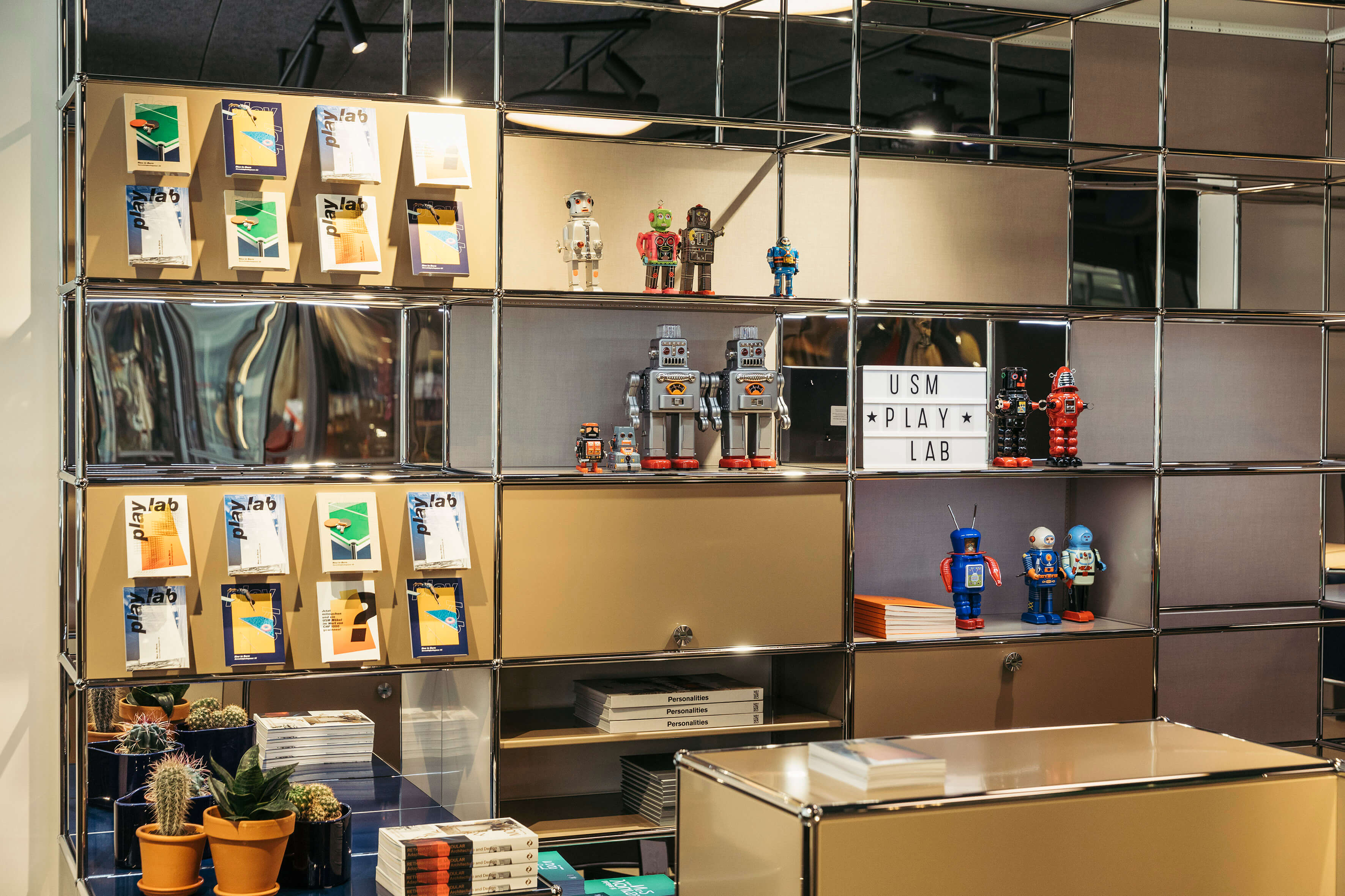 USM Regalsystem mit verchromtem Rahmen und goldenen Paneelen, bestückt mit Play Lab Broschüren, kleinen Roboterfiguren und Leuchtbox mit Schriftzug „USM PLAY LAB“.