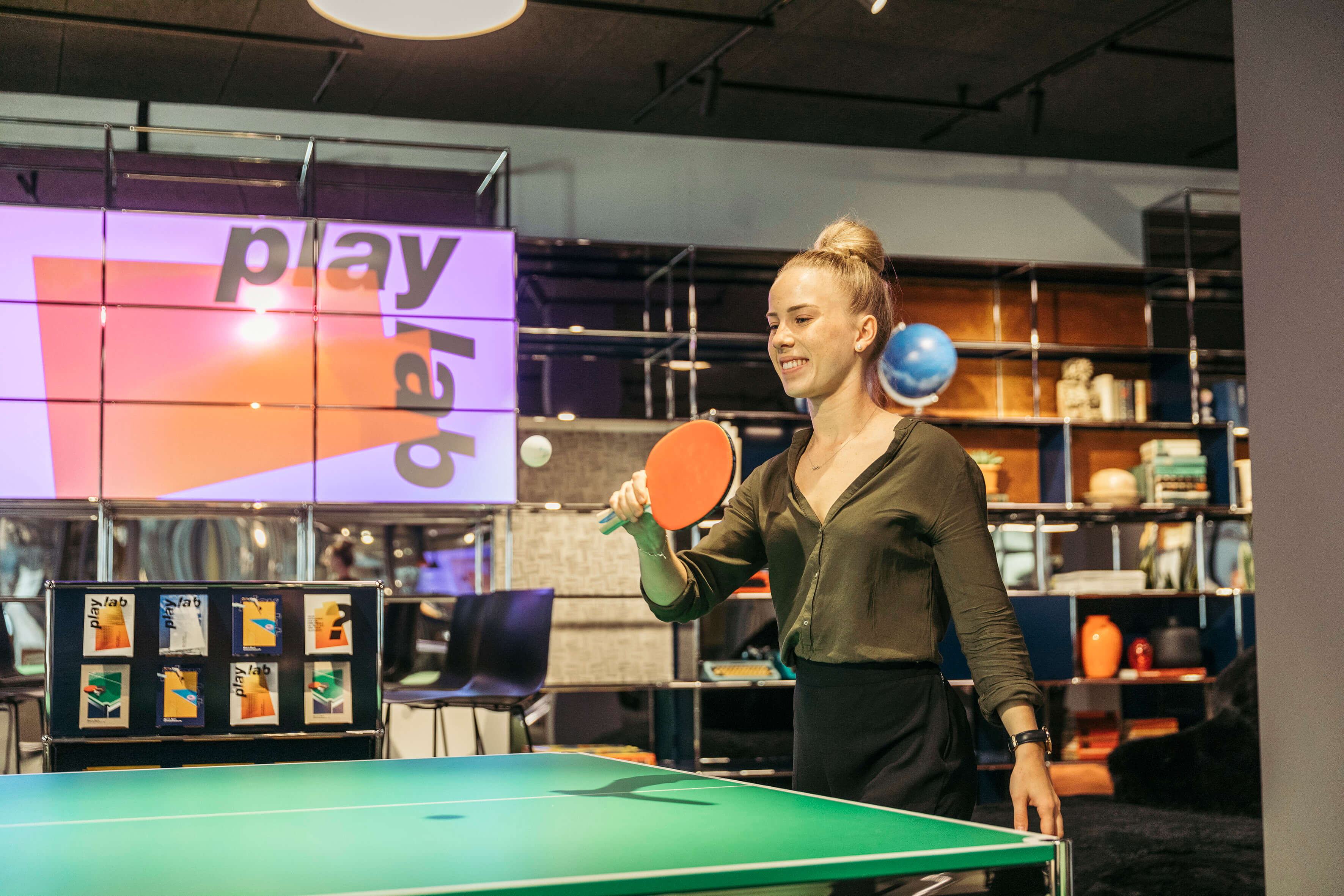 Person spielt Tischtennis an einem grünen Tisch, Ball in der Luft, im Hintergrund USM Regale und beleuchtetes „play lab“ Branding.