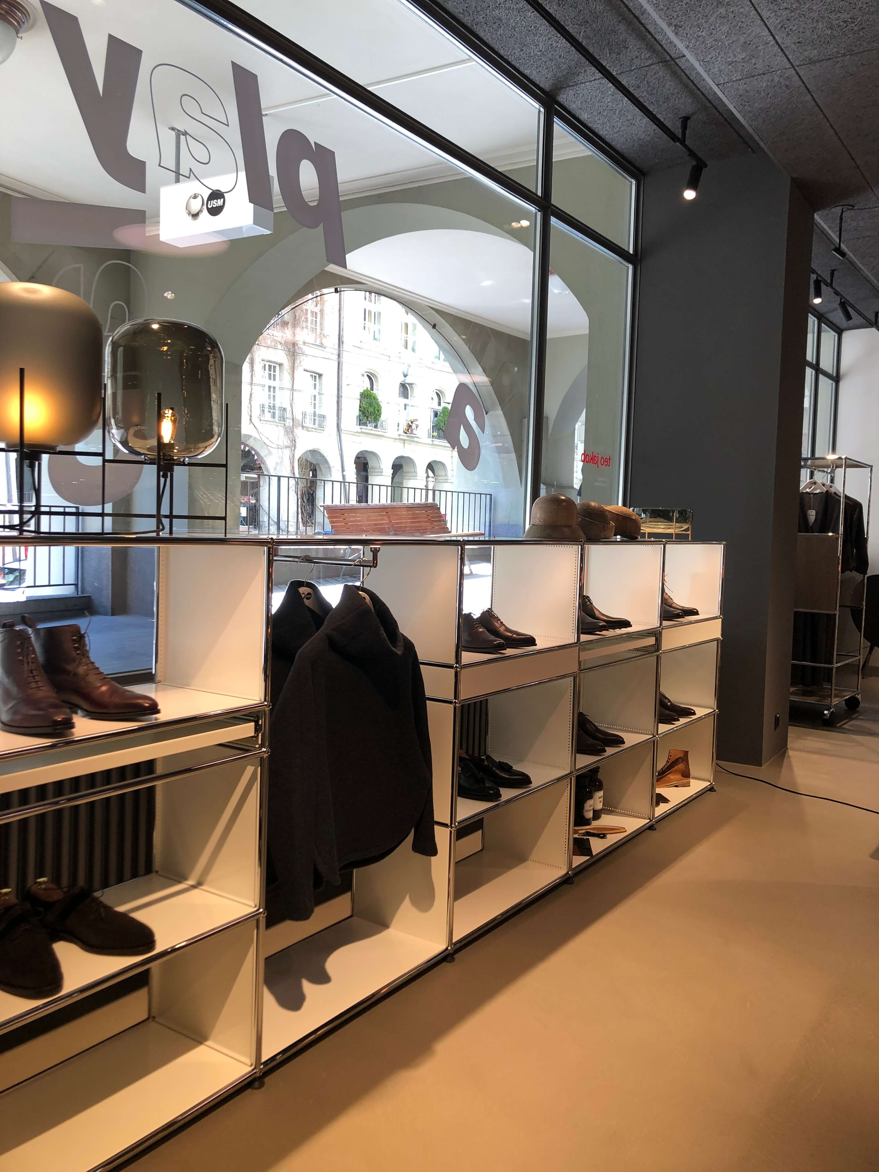 Showroom mit modularen Regalen und offenen Fächern, bestückt mit Schuhen und Jacken. Grosse Fensterfront mit Blick auf Arkaden und Strassenfassaden, beleuchtet durch Schienenstrahler und eine Tischleuchte.