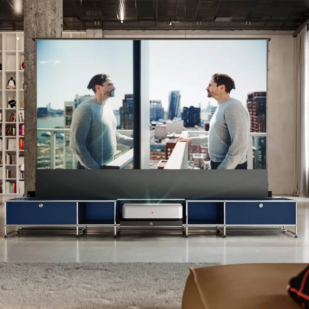 Innenraum mit grosser Projektionsleinwand. Ein Kurzdistanzprojektor steht auf einem blauen USM Haller Lowboard und projiziert eine Szene mit einem Mann auf einem Balkon vor Stadtsilhouette. Loftambiente mit Sichtbeton, Bücherregal und viel Tageslicht.