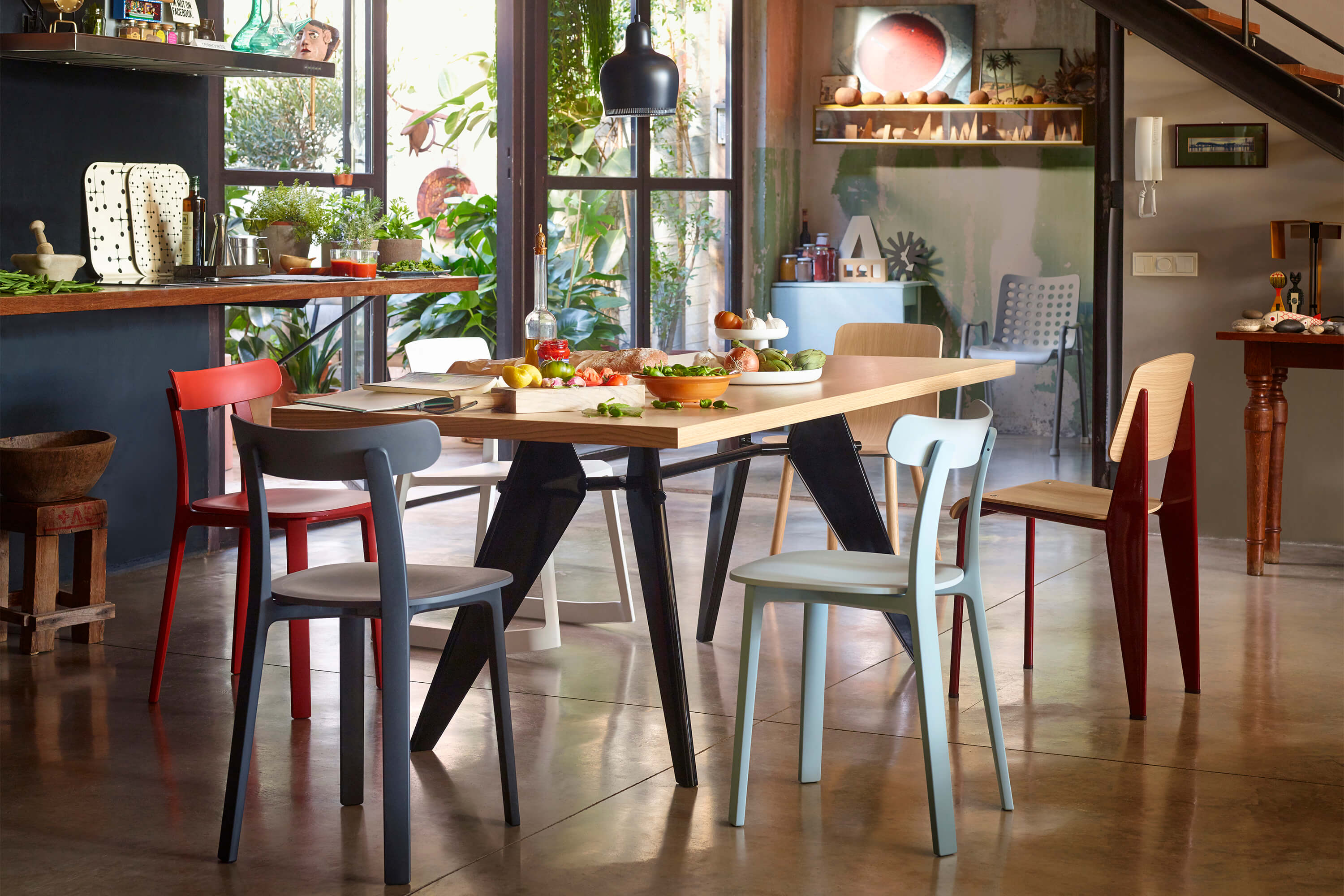 Lebendiger Essbereich mit grossem Holztisch auf poliertem Betonboden. Rundum stehen bunte Stühle in Rot, Grau, Hellblau und Holz. Auf dem Tisch liegen Brot, Gemüse und Geschirr für ein gemeinsames Essen. Im Hintergrund sorgen grosse Fenster, viele Pflanzen und offene Regale für Loft Atmosphäre. Eine schwarze Pendelleuchte hängt über dem Tisch, rechts führt eine Treppe nach oben.