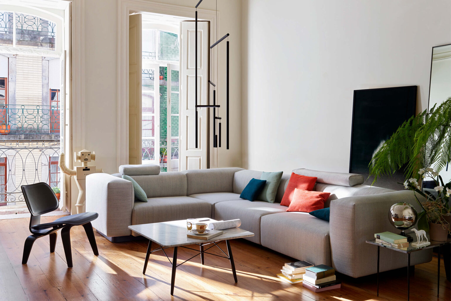 Helles Wohnzimmer mit grauem Ecksofa und farbigen Kissen in Türkis und Rot. Vor dem Sofa steht ein niedriger Couchtisch mit Tassen und Büchern. Links führen hohe Fenstertüren auf einen Balkon, nebenan steht ein schwarzer Stuhl und eine skulpturale Stehleuchte. Rechts ein Beistelltisch mit Pflanzen und Deko, dazu ein grosses Bild an der Wand. Warmes Tageslicht fällt auf den Holzboden.