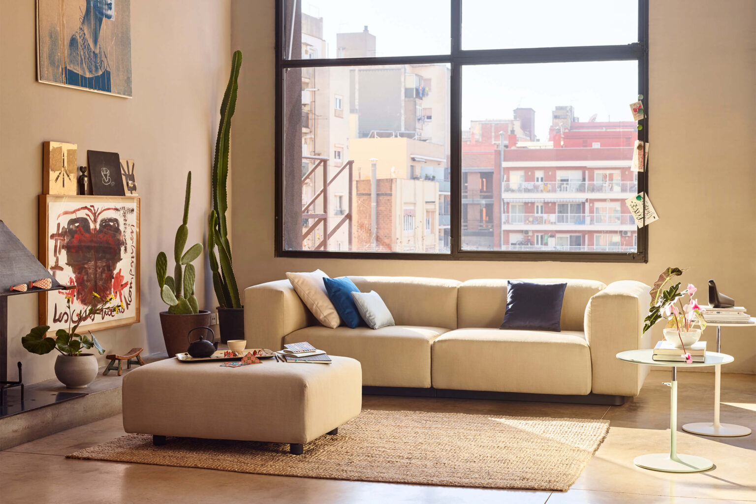 Heller Wohnraum mit beigem Sofa vor grossem Industrie Fenster mit Stadtblick. Links stehen hohe Kakteen neben gerahmter Kunst und Bildern auf dem Boden. Vor dem Sofa liegt ein Pouf auf Jute Teppich, rechts ein runder Beistelltisch mit Vase und Büchern. Warmes Tageslicht prägt die Szene.