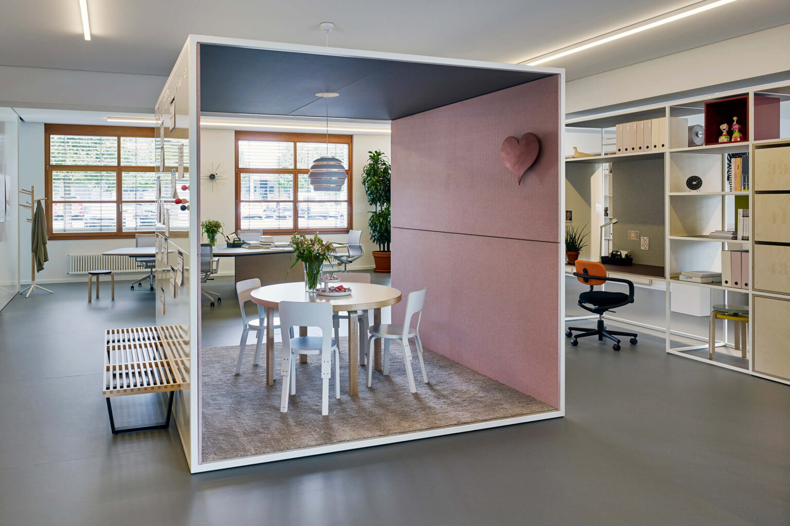 Offenes Büro mit Raum-in-Raum Box als Besprechungszone. In der Box stehen ein runder Tisch mit weissen Stühlen auf Teppich, die Innenwand ist in Rosé gepolstert und mit einem Herzobjekt akzentuiert. Rundherum sind Arbeitsplätze, Regale, Pflanzen und grosse Fenster sichtbar. Lineare Deckenleuchten und ein Hängelicht über dem Tisch strukturieren den Raum.