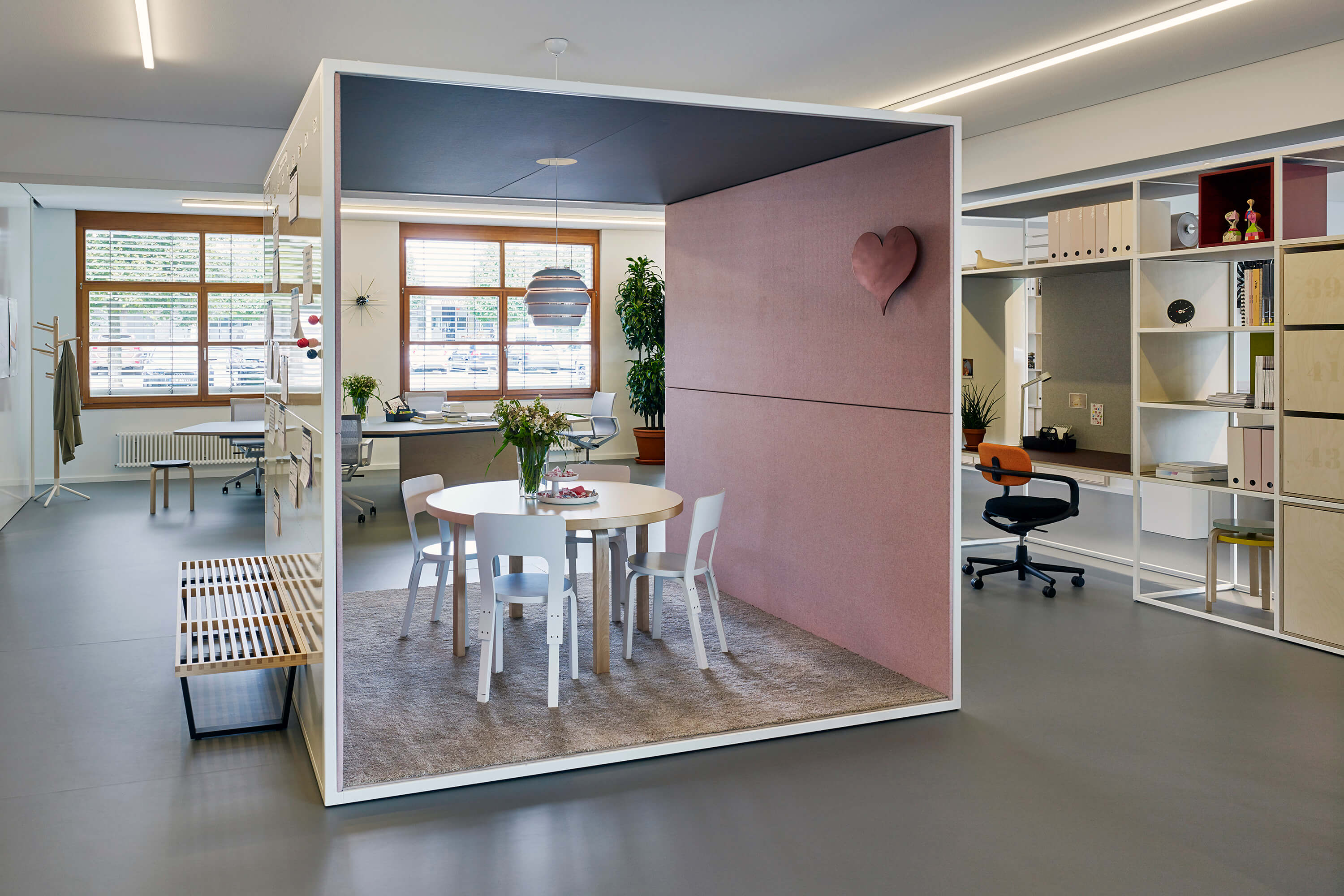 Offenes Büro mit Raum-in-Raum Box als Besprechungszone. In der Box stehen ein runder Tisch mit weissen Stühlen auf Teppich, die Innenwand ist in Rosé gepolstert und mit einem Herzobjekt akzentuiert. Rundherum sind Arbeitsplätze, Regale, Pflanzen und grosse Fenster sichtbar. Lineare Deckenleuchten und ein Hängelicht über dem Tisch strukturieren den Raum.
