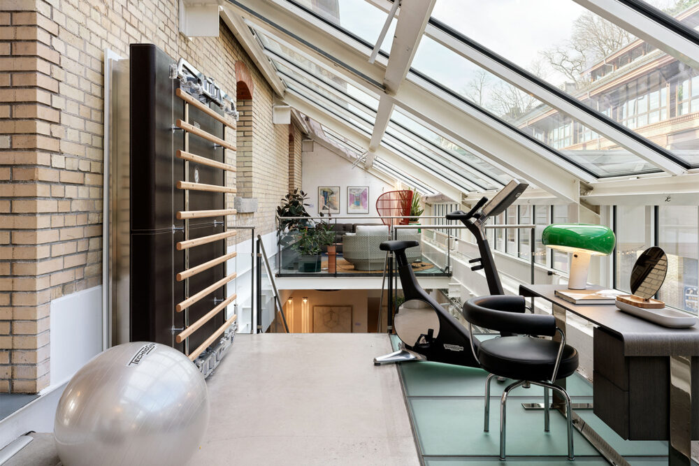 Heller Fitnessbereich unter Glasdach. Sprossenwand, Ergometer und Gymnastikball neben einem Schreibtisch mit grüner Bankerslampe. Backsteinwände, Galerieebene mit Lounge und Pflanzen im Hintergrund.