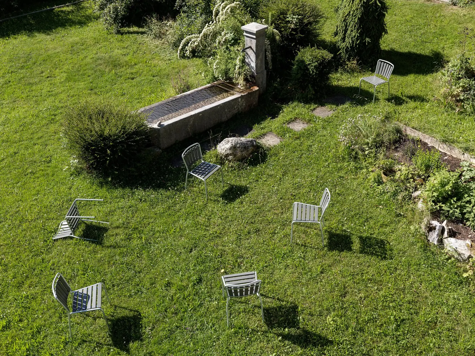 Gartenrasen mit sechs verteilten Metallstühlen in der Sonne, daneben ein steinernes Brunnenbecken, Trittsteine und dichte Bepflanzung.